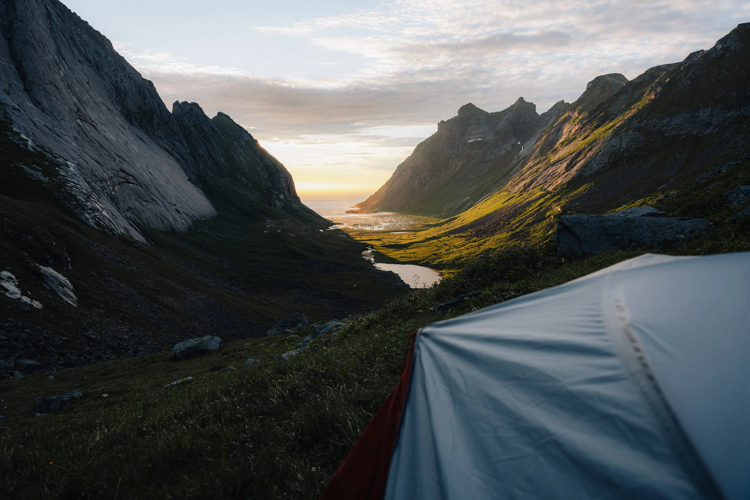 A scenic view of a valley between tall, rugged mountains during sunset or sunrise, with a tent in the foreground.
