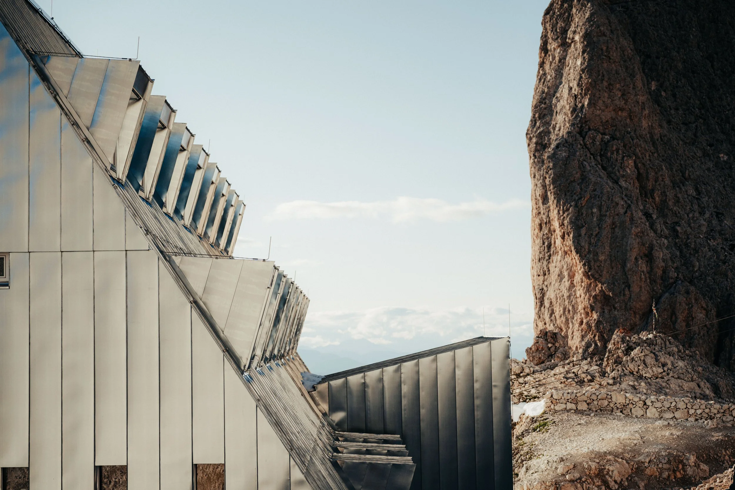 Modern building with metallic panels on the left side and a large rocky mountain on the right side under a partly cloudy sky.
