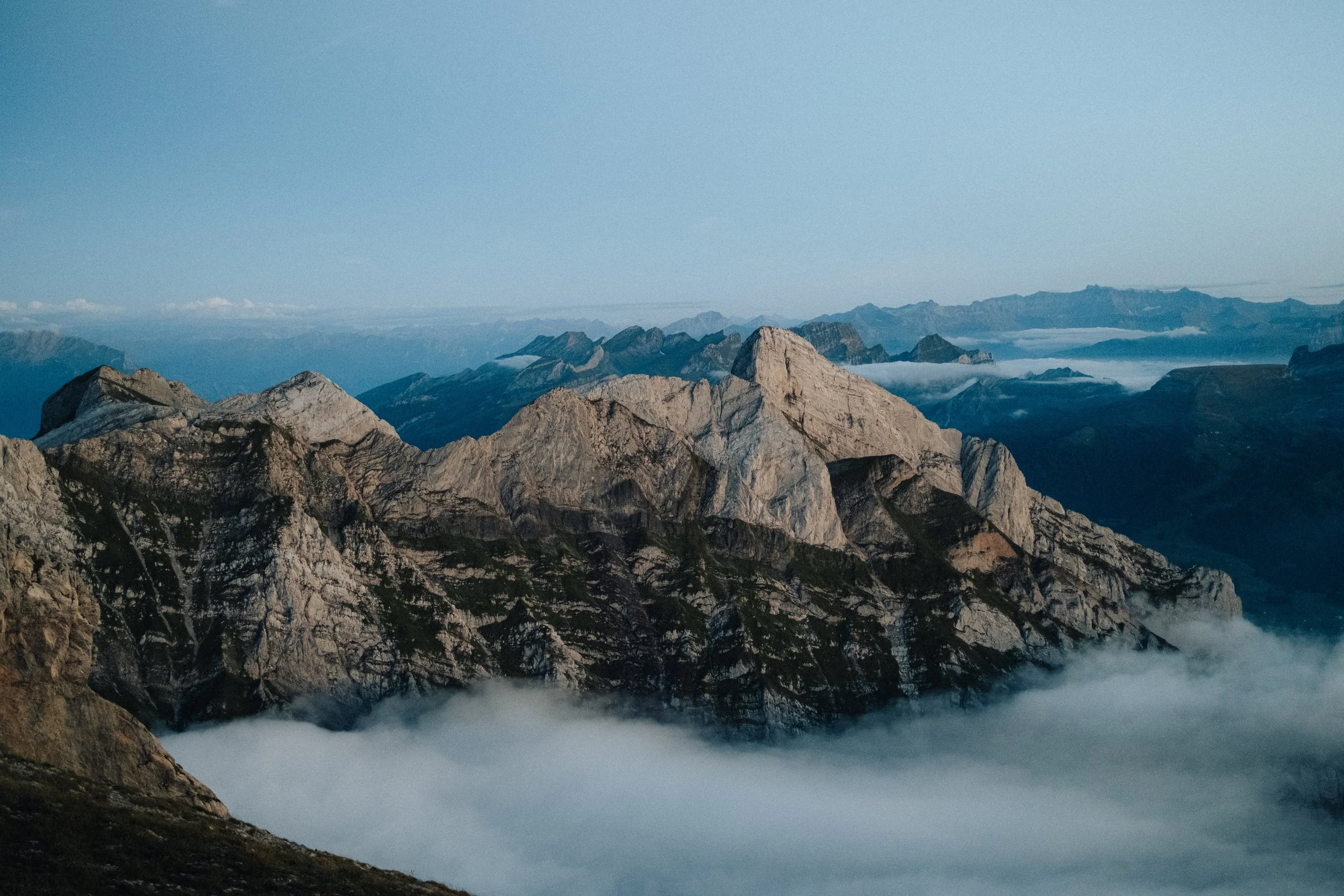 A scenic mountain landscape with rocky peaks and a layer of clouds below in the foreground, with distant mountains and a clear sky in the background.