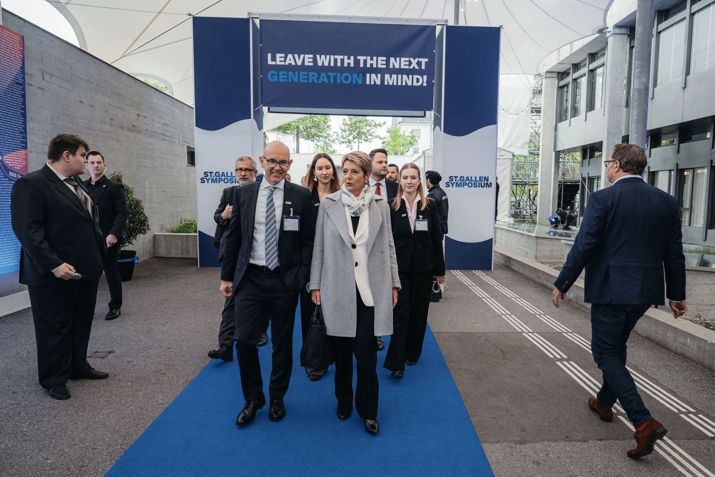 Group of professionally dressed people walking under a sign that reads, 'Leave with the next generation in mind!' at the ST. GALLEN SYMPOSIUM conference.