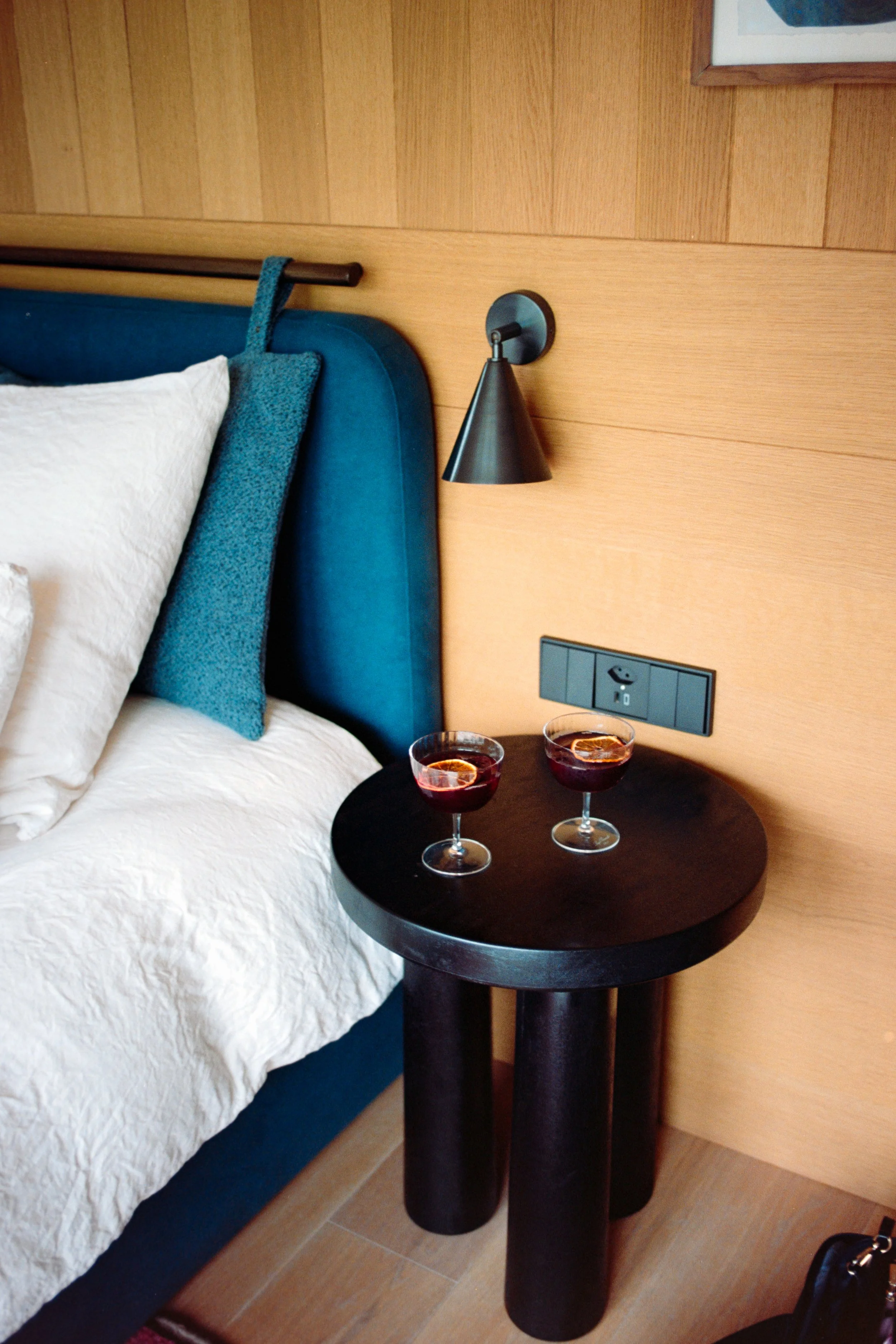 A bedroom with a bed that has white bedding and a blue upholstered headboard, a small black side table with two glasses of red wine, black wall-mounted reading light, and a wooden wall with a power outlet.