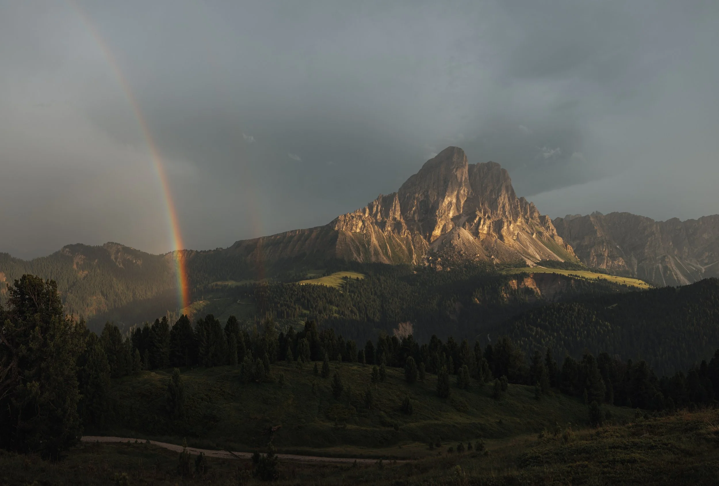 Mountain landscape with dark clouds, a rainbow, and lush green trees in the foreground.