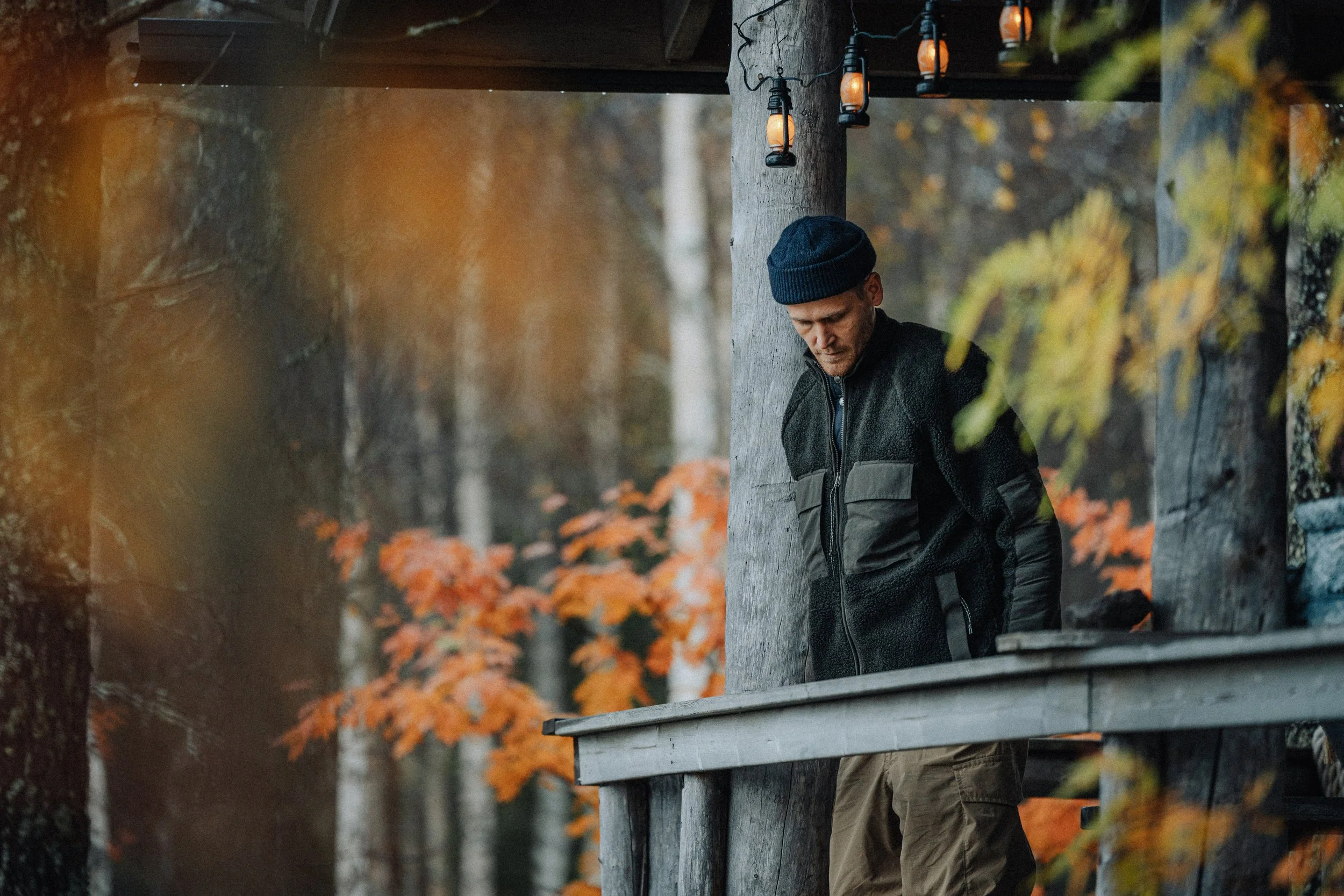A man with a dark beanie hat and black jacket standing on a wooden balcony surrounded by fall leaves.