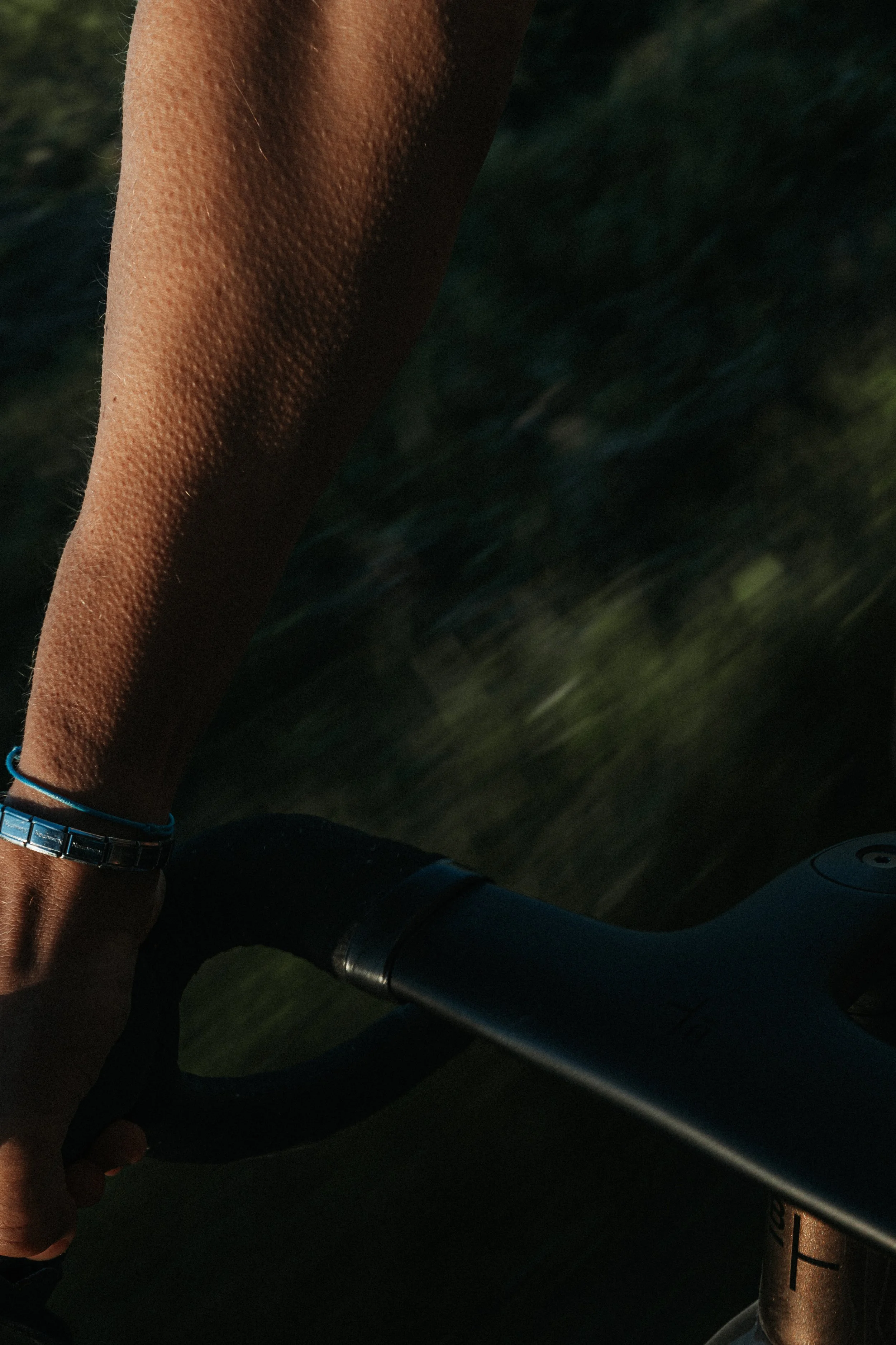 A person's arm with a blue bracelet, gripping a bicycle handlebar, with blurred green scenery in the background at sunset.