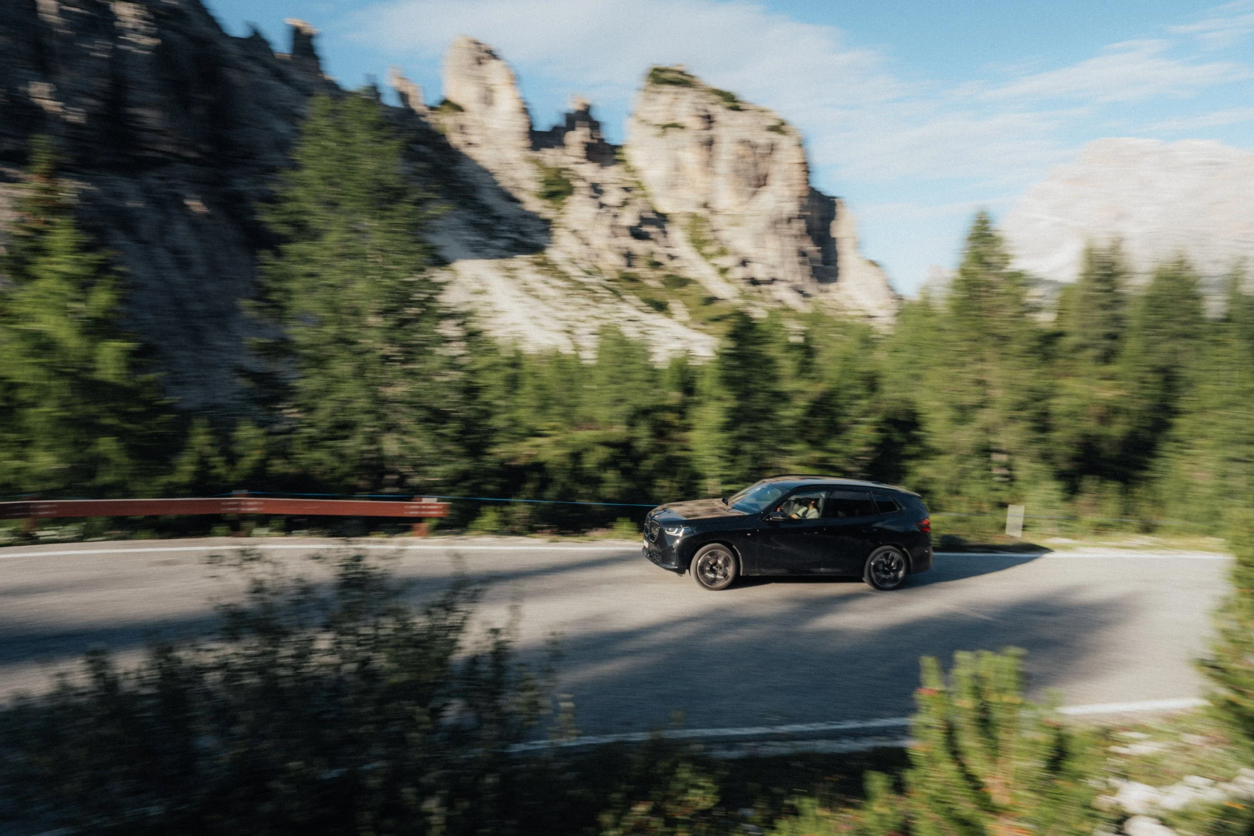 A black SUV driving on a scenic mountain road with rocky cliffs and green trees in the background.