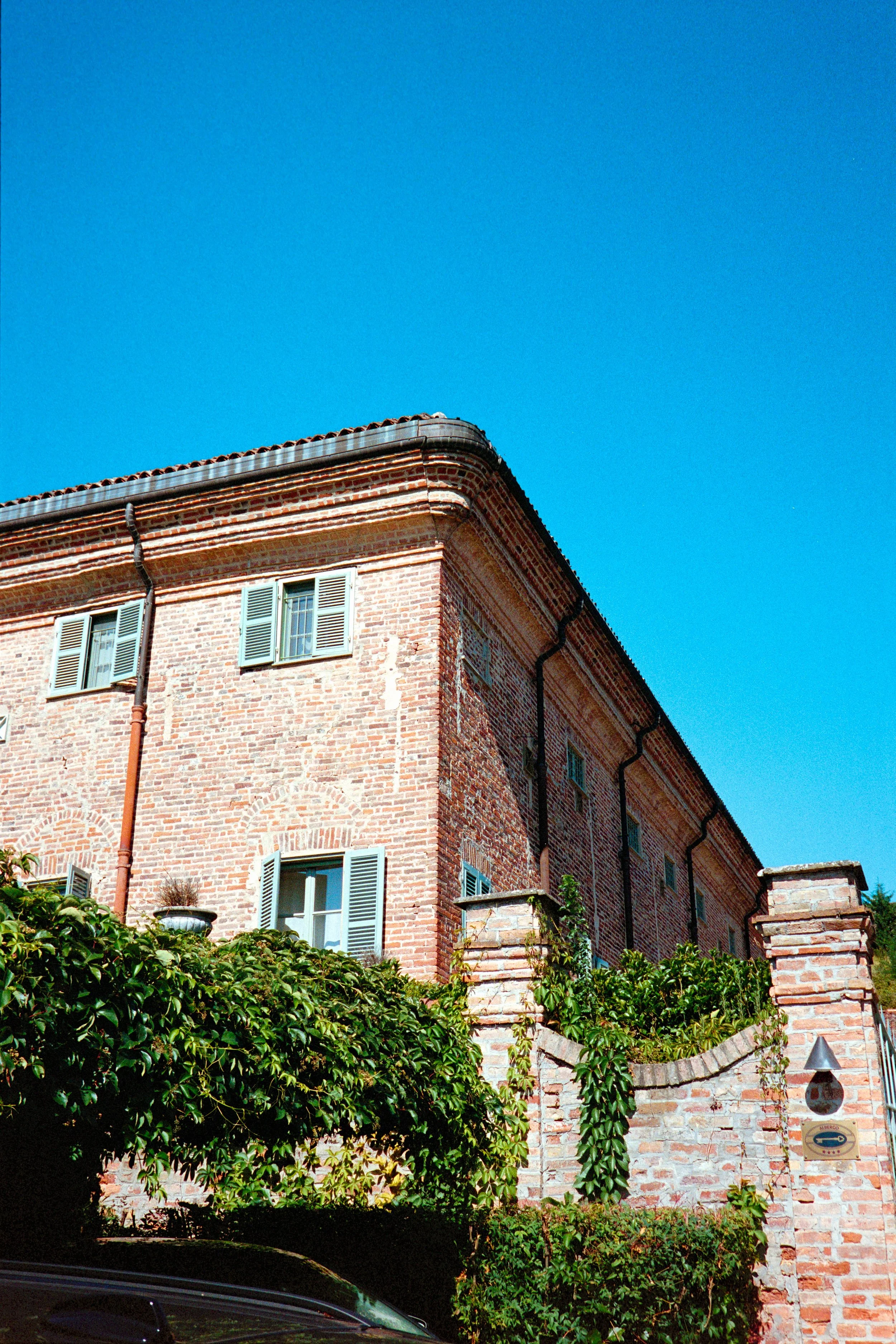 A brick building with windows and shutters, surrounded by greenery, under a clear blue sky.