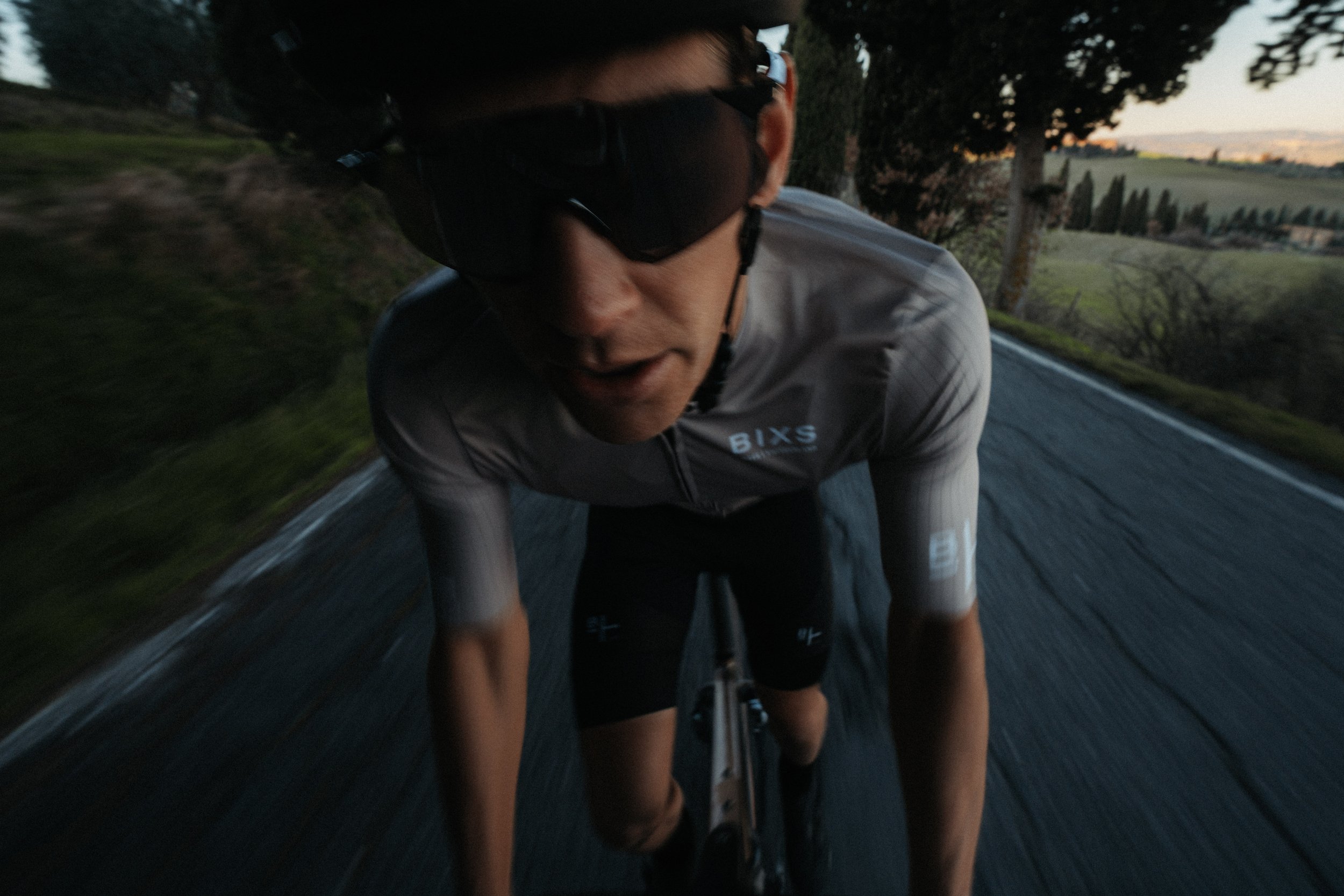 A person riding a bicycle on a rural road during dusk, wearing sunglasses and a gray cycling jersey.