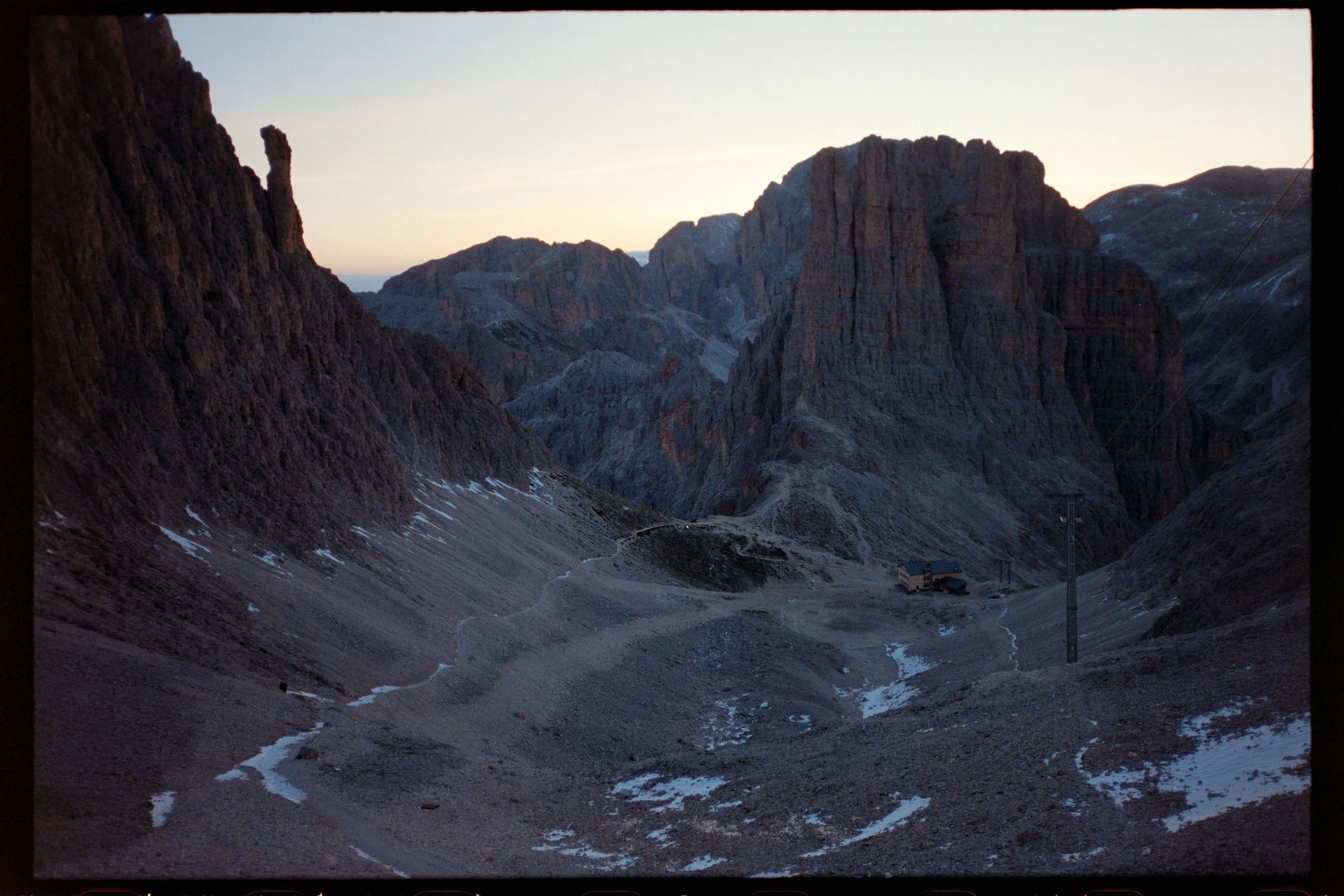 Mountain landscape with steep rocky cliffs, a narrow trail, patches of snow, and a small building at the base, during dusk or dawn.