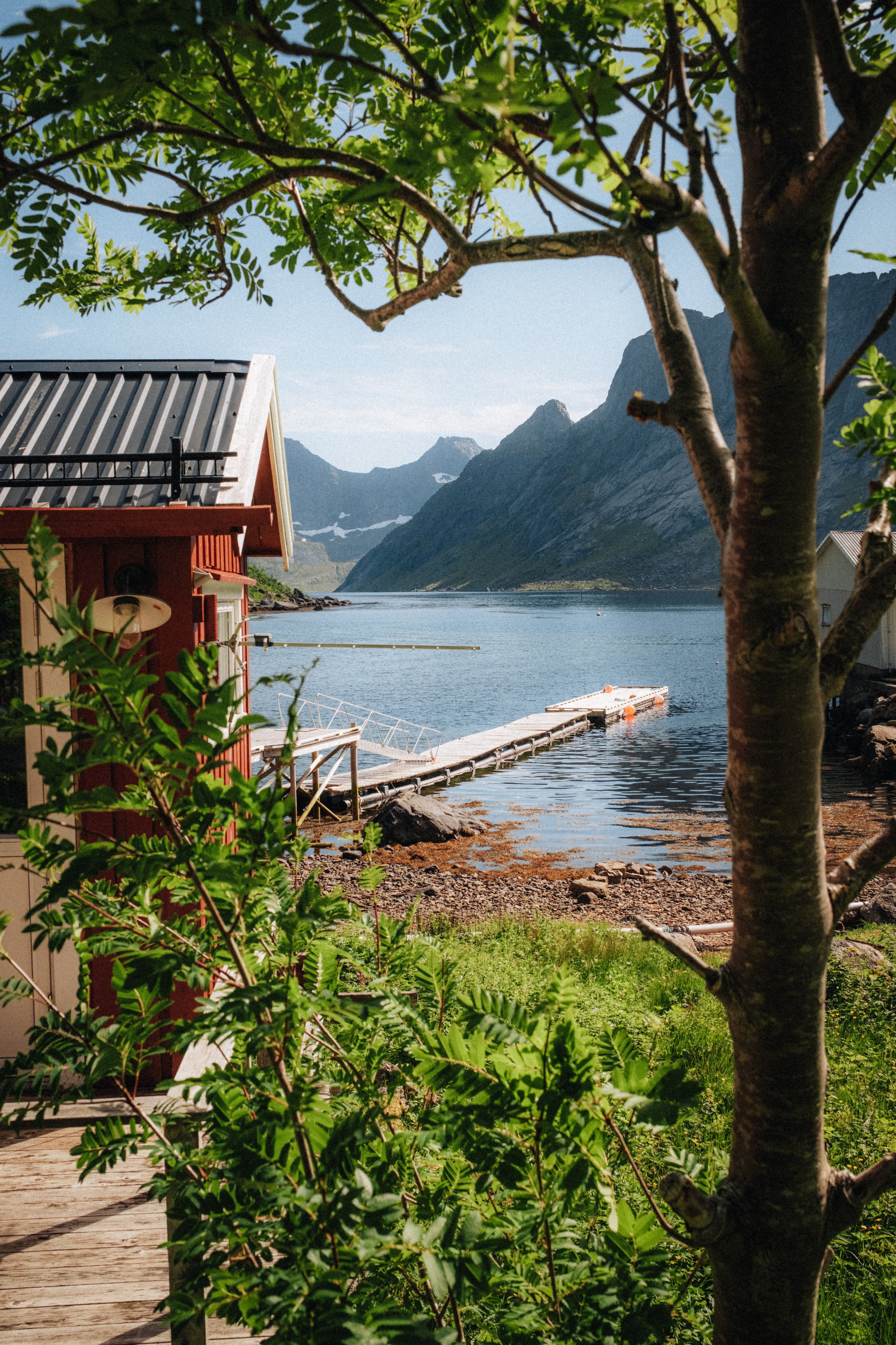 Scenic view of a wooden dock leading into a calm body of water, surrounded by green mountains with snow patches, with a red cabin partially visible on the left, and leafy green trees in the foreground.
