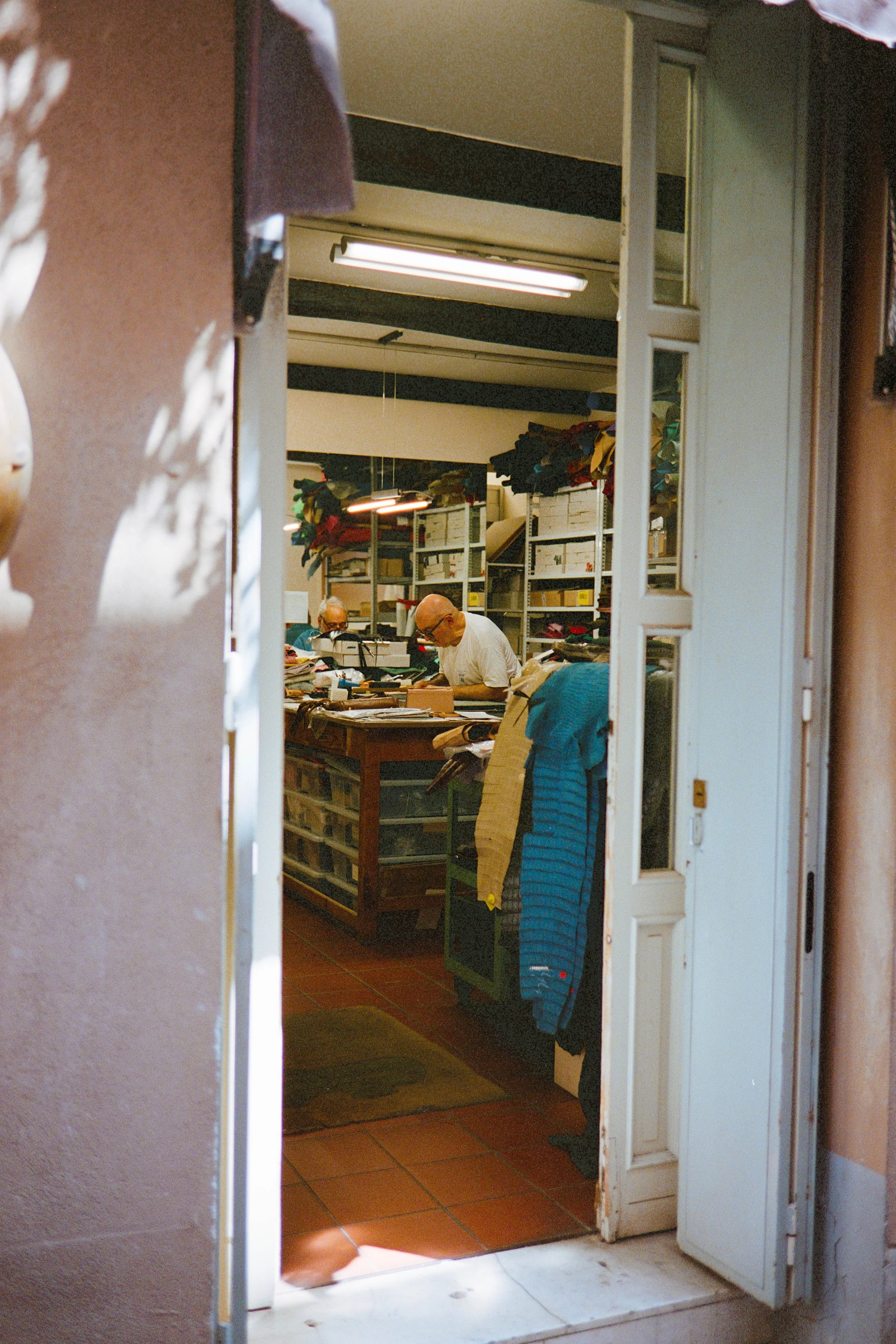 View of a workshop with two men working at a cluttered table, surrounded by shelves and boxes filled with fabric and materials, seen through an open door.
