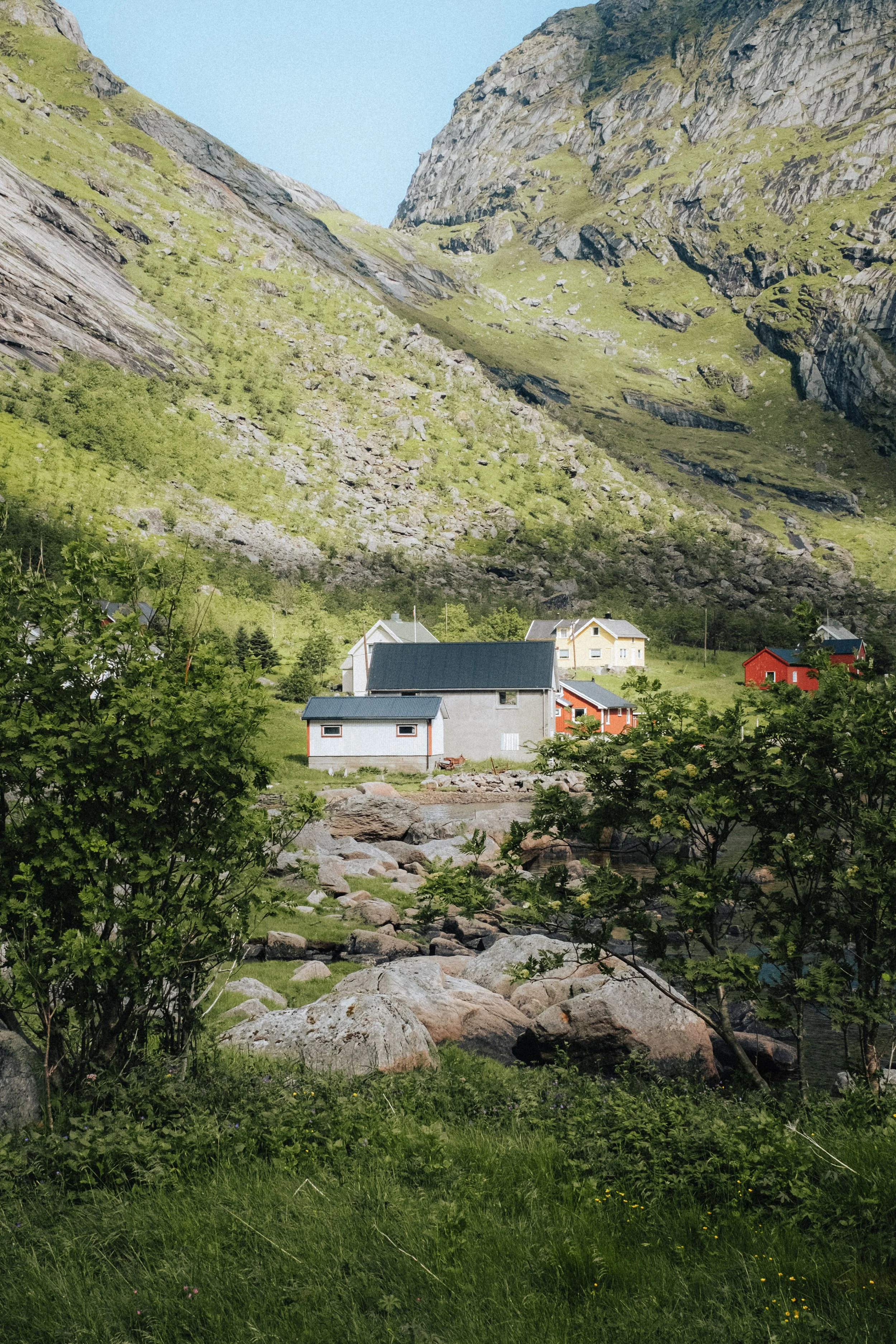 A scenic view of a small village at the foot of a steep, green mountain with rocks and a stream in the foreground.