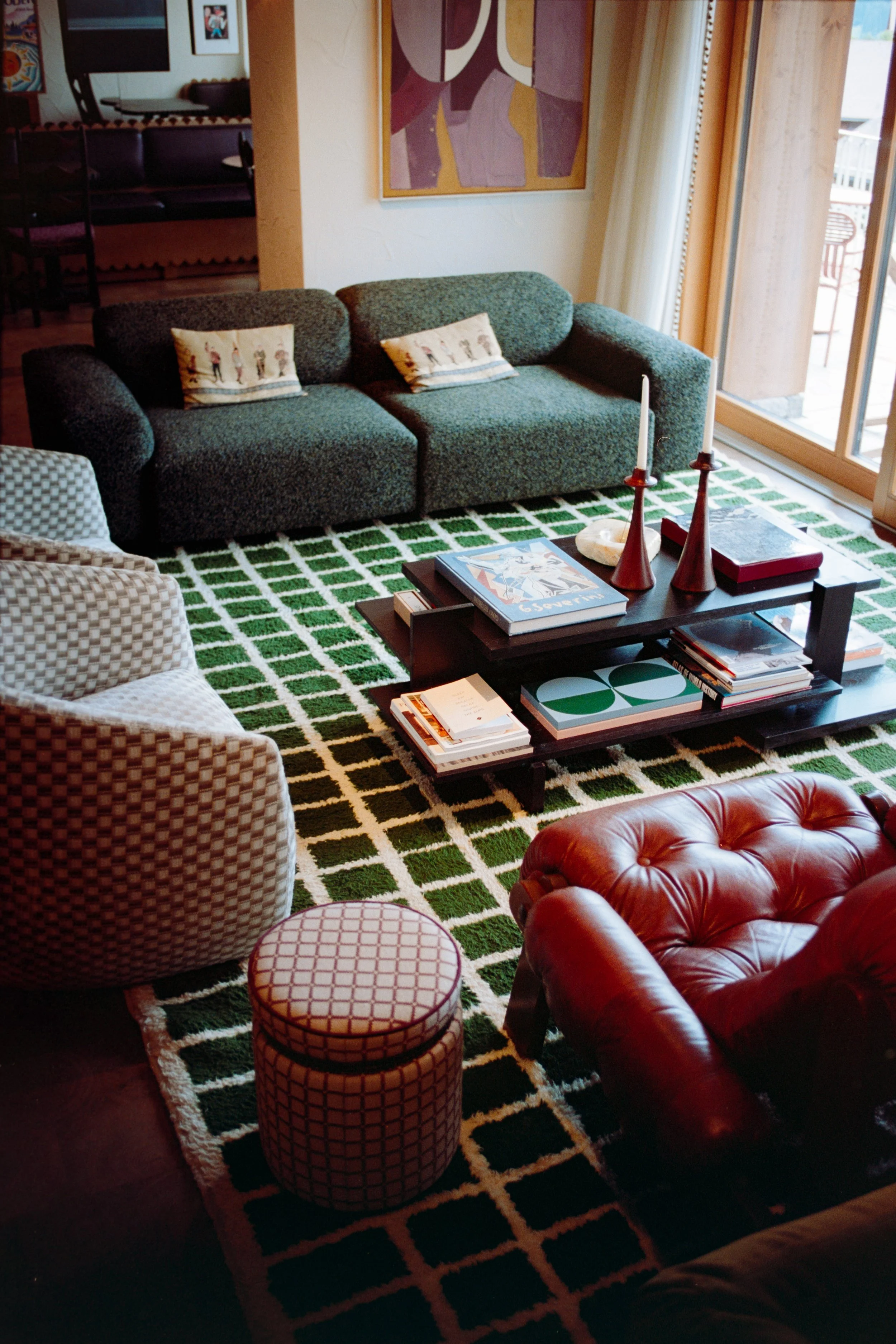 A cozy living room with a green sofa, patterned armchair, red leather armchair, patterned ottoman, black coffee table with books and candles, a colorful abstract painting on the wall, and glass sliding doors leading to a balcony.
