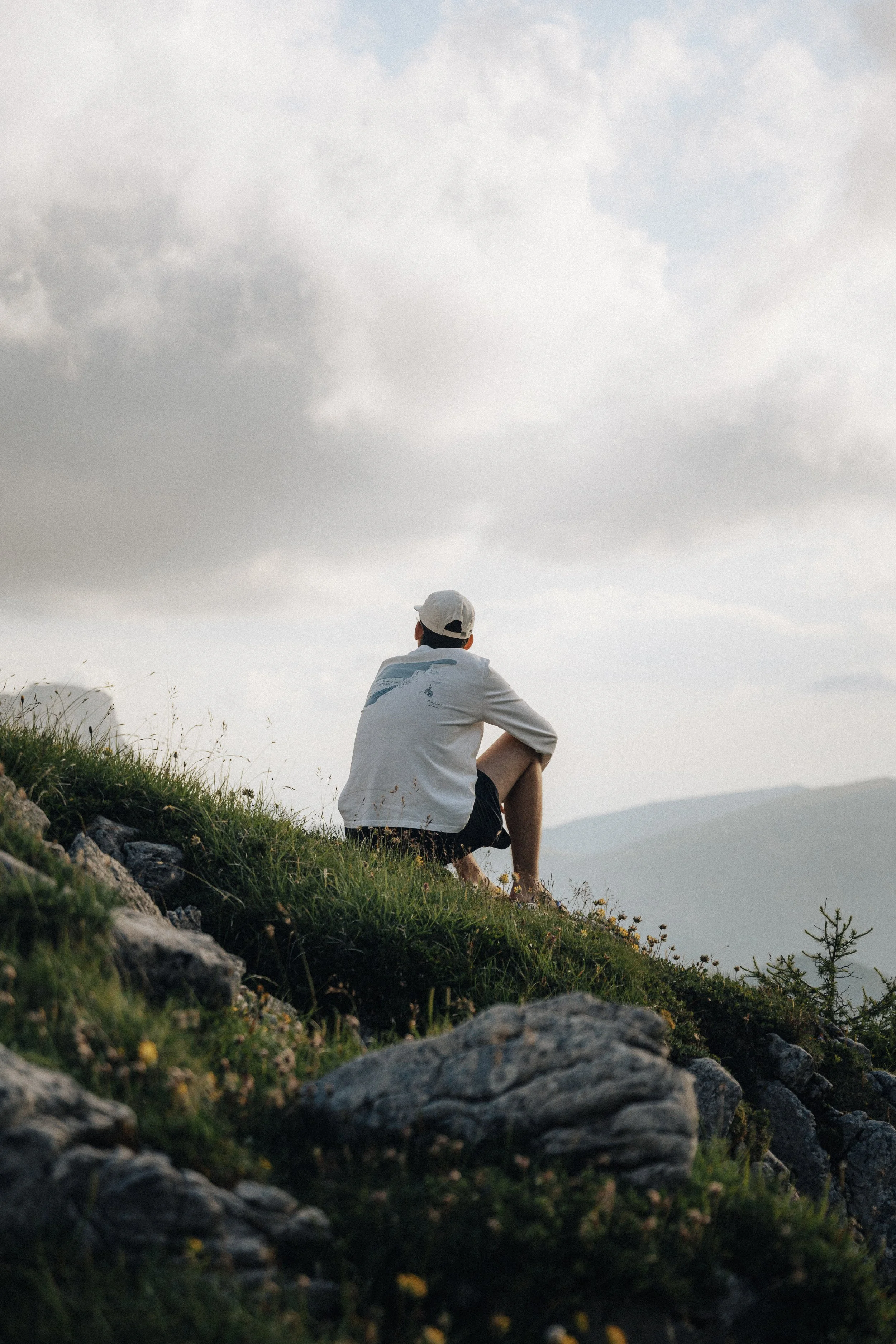 A person sitting on a grassy hillside overlooking distant mountains, wearing a white jacket, shorts, and a white cap, gazing at the cloudy sky.