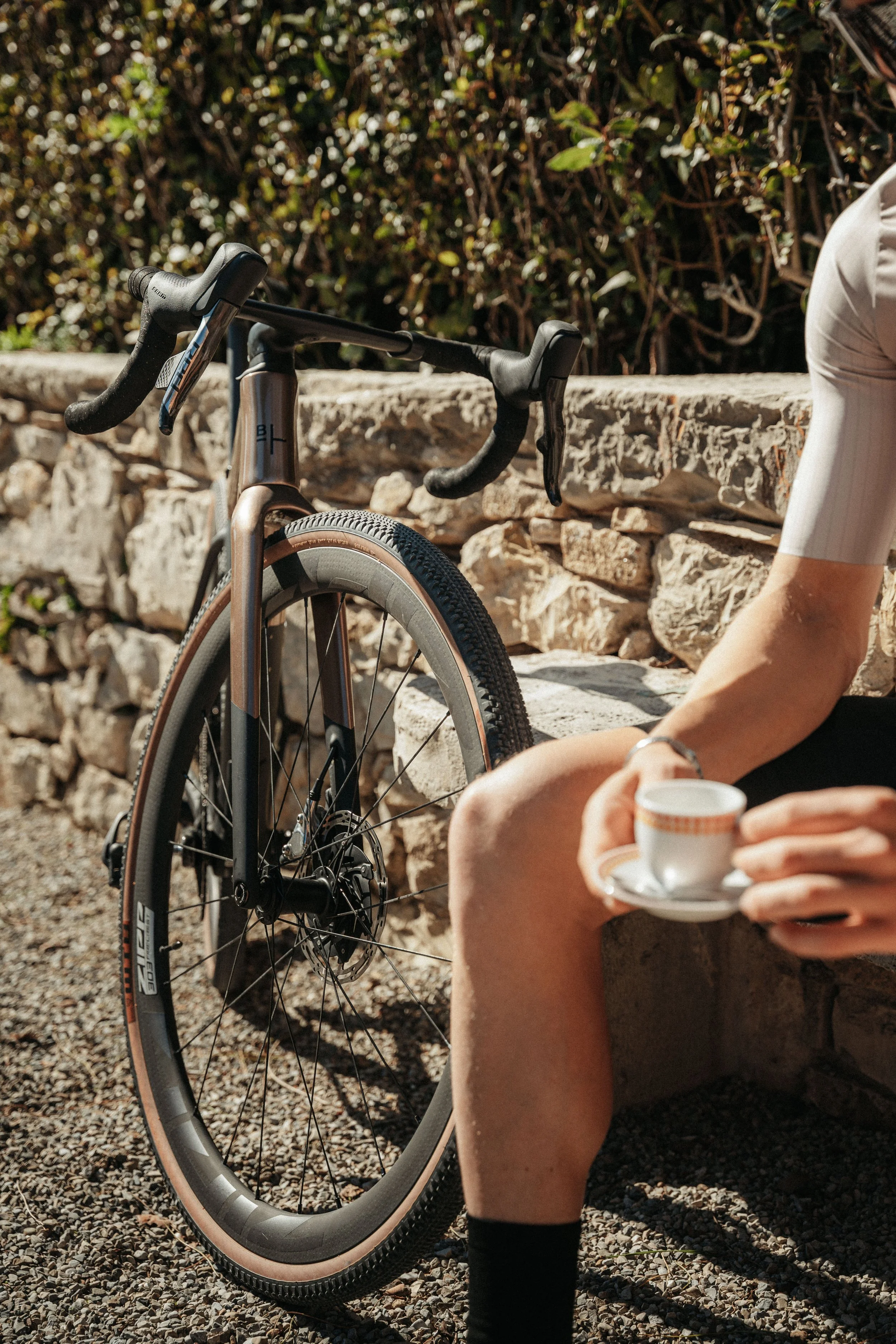 A person sitting on a stone bench, holding a small teacup with a saucer in hand, with a bicycle leaning against the stone wall beside them. The scene appears to be outdoors with a stone wall and some greenery in the background.