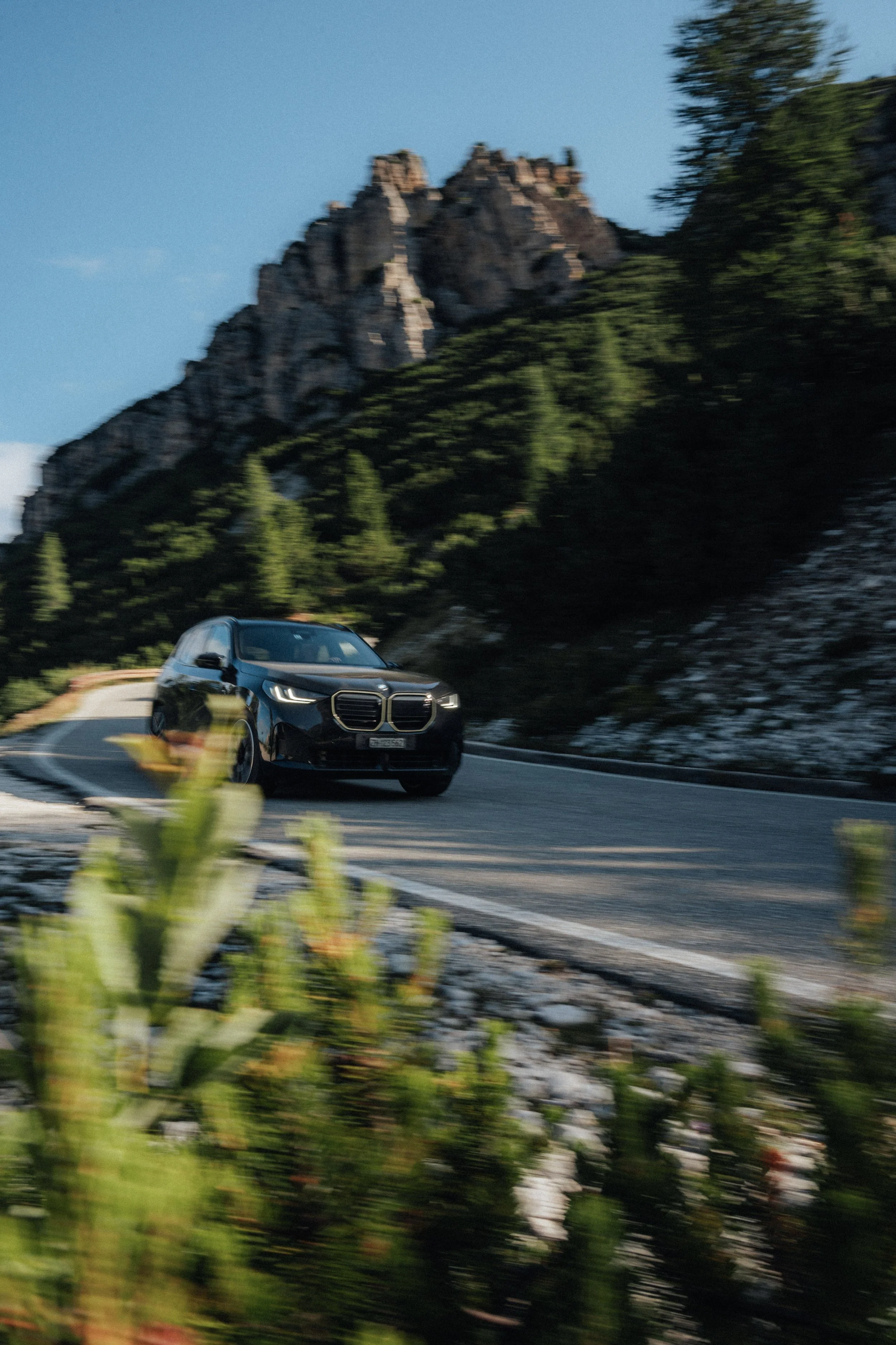 A black car driving on a mountain road with rocky cliffs and greenery in the background.