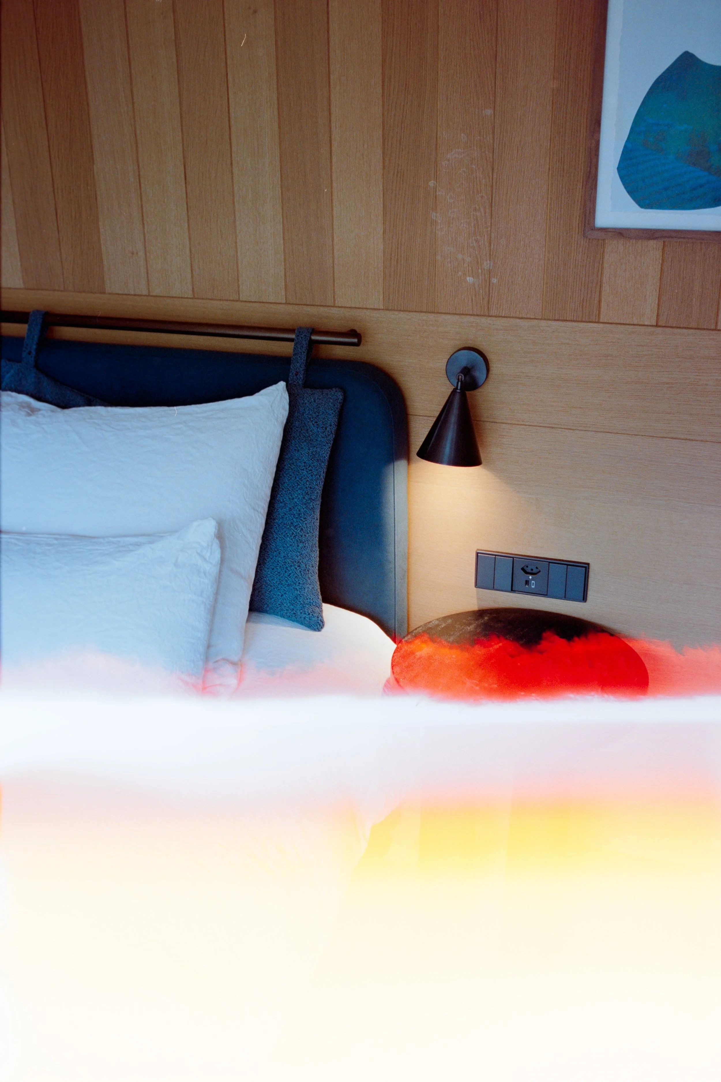 A bedroom with a bed featuring white bedding and pillows, a wooden headboard, a wall-mounted black lamp, and a wooden wall with a framed artwork.