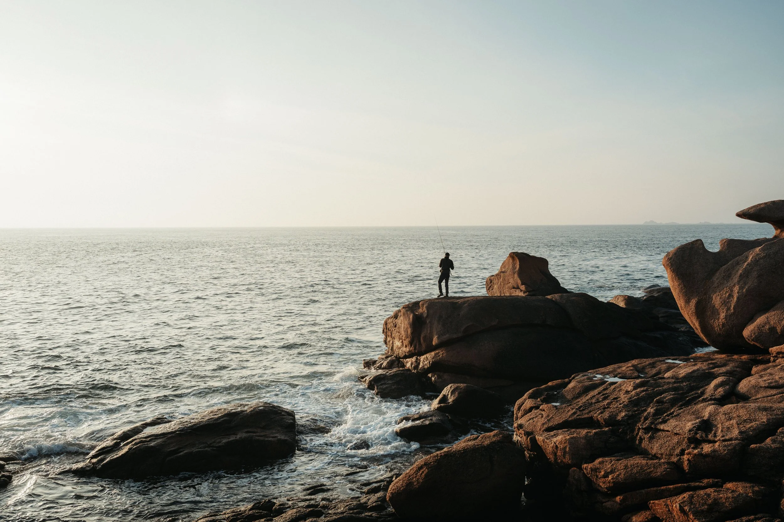 A person standing on rocks by the sea, fishing, with the ocean and sky in the background.