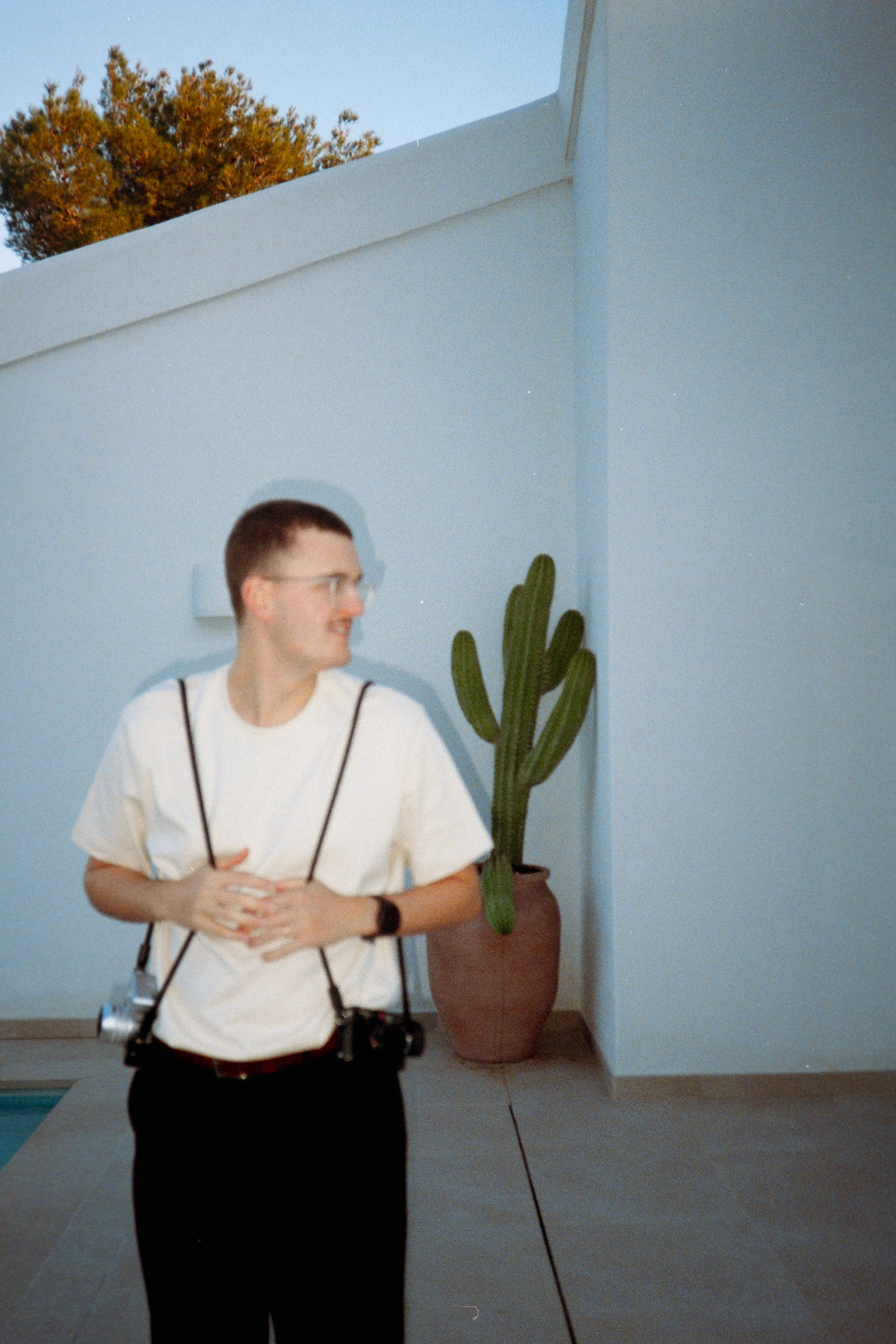 A person with short dark hair and glasses, dressed in a white t-shirt and black pants, standing outdoors near a light blue wall with a large potted cactus plant.