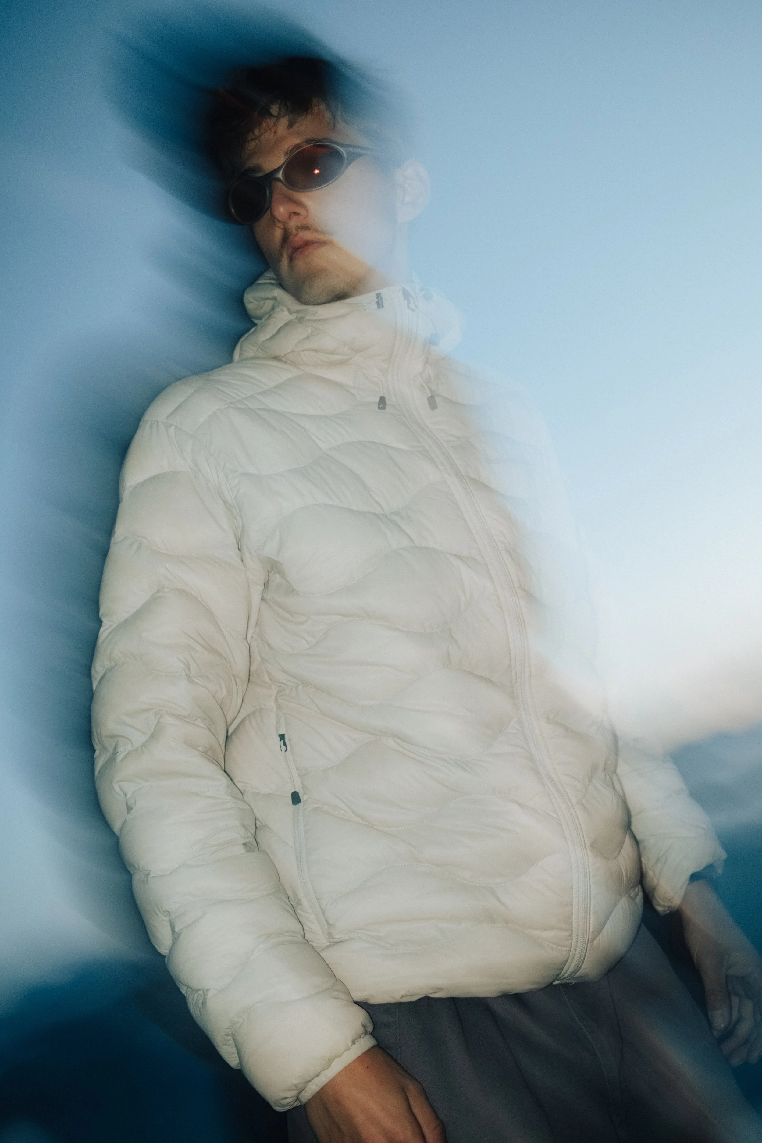 A young man wearing sunglasses and a white puffy jacket leaning against a blue background, with motion blur creating a sense of movement.