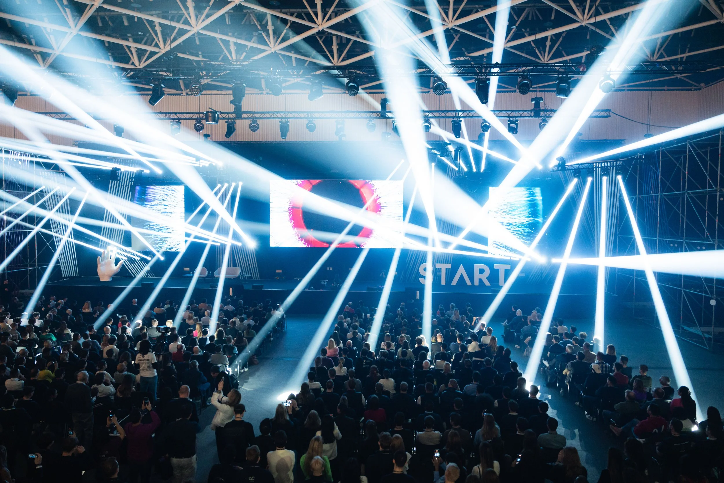 Large audience at a concert or event with bright white stage lights, LED screens showing visuals, and a stage setup with a "START" sign.