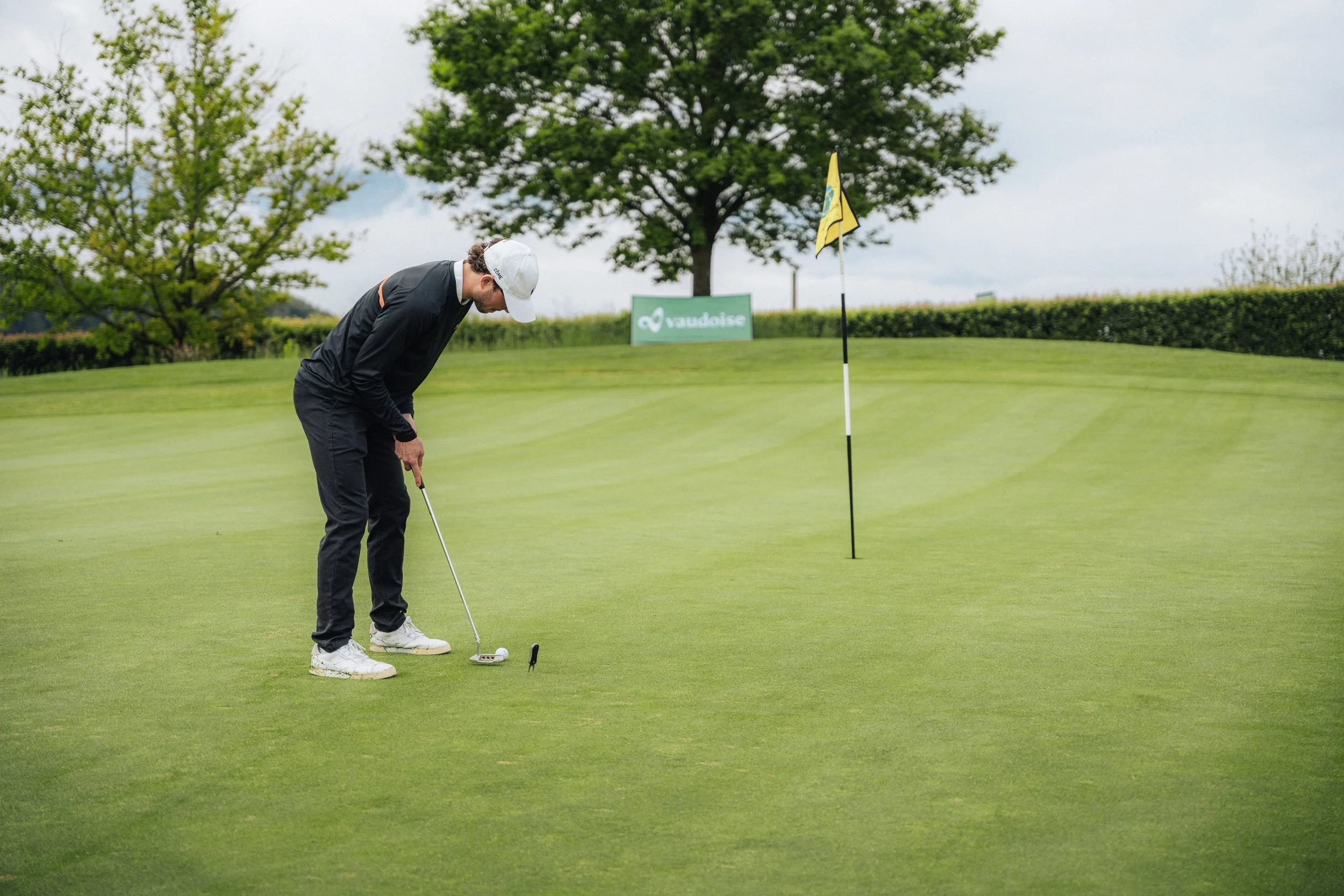 A man in black golf attire is putting on a golf green, with a yellow flagstick marking the hole, a sign with the word "Vaudoise" is visible in the background, along with trees and bushes.