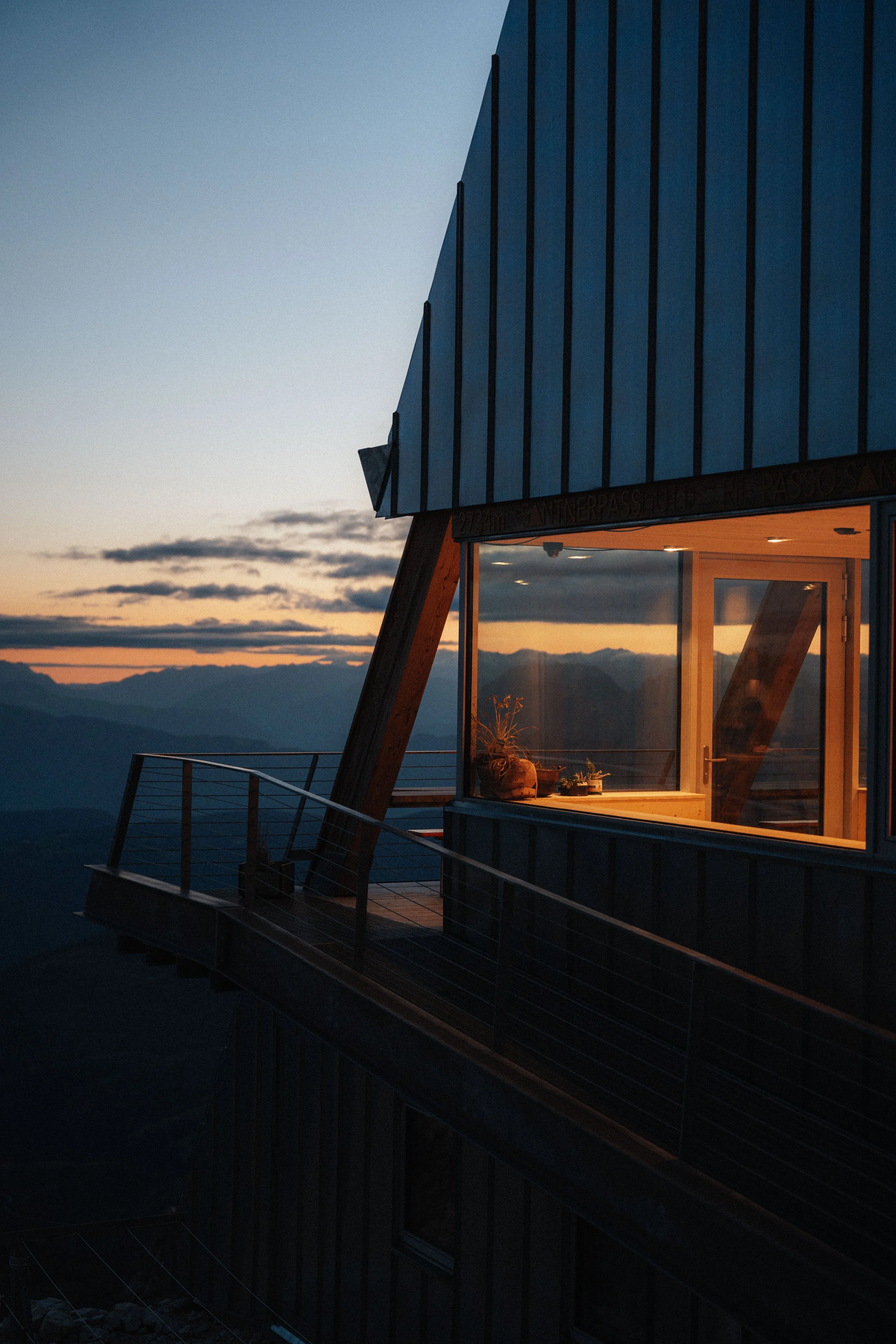 A modern mountain building with large windows and a balcony during sunset, with mountain ranges in the background.