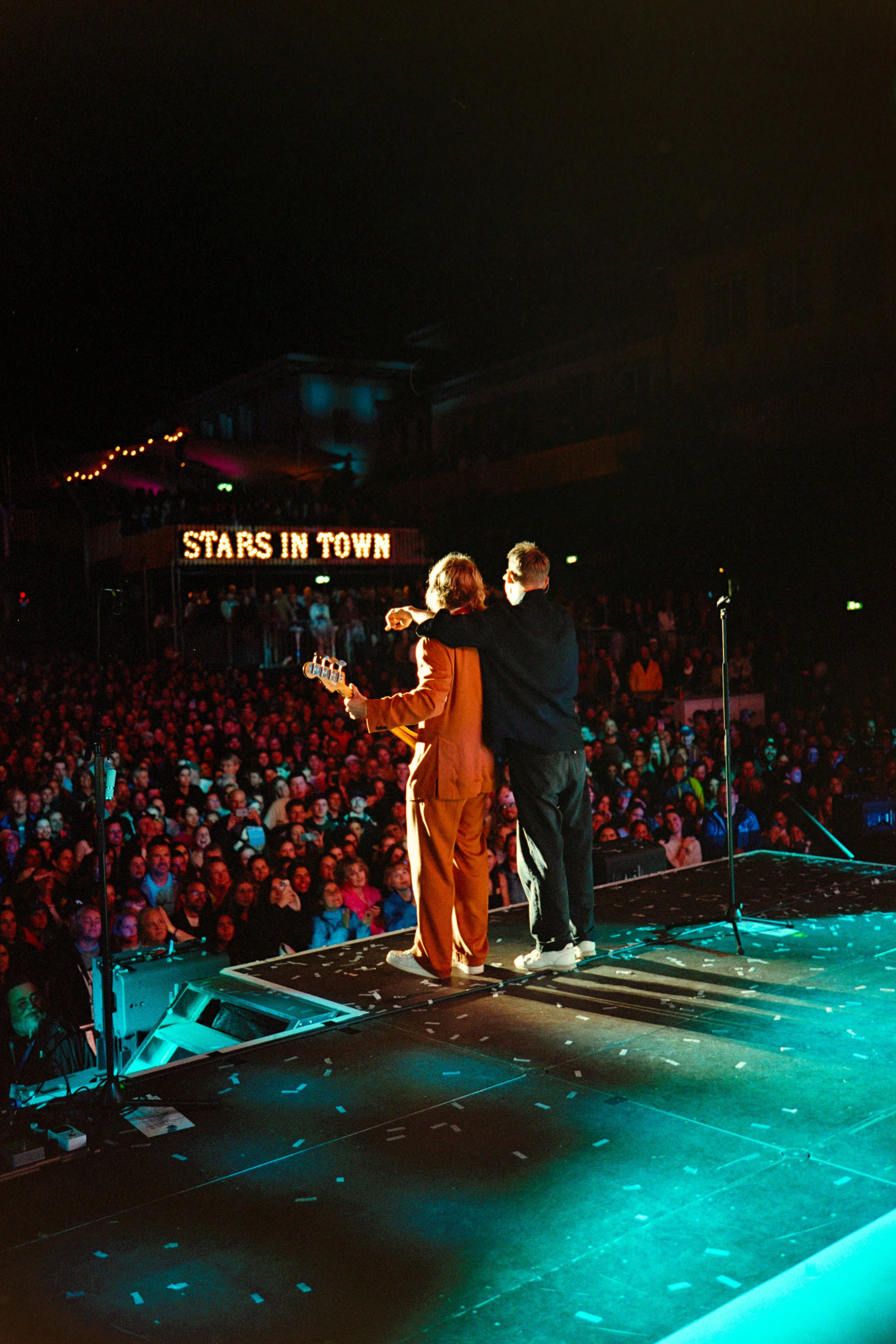 Two performers on stage in front of a large audience during a concert, with a sign in the background reading 'Stars in Town'.