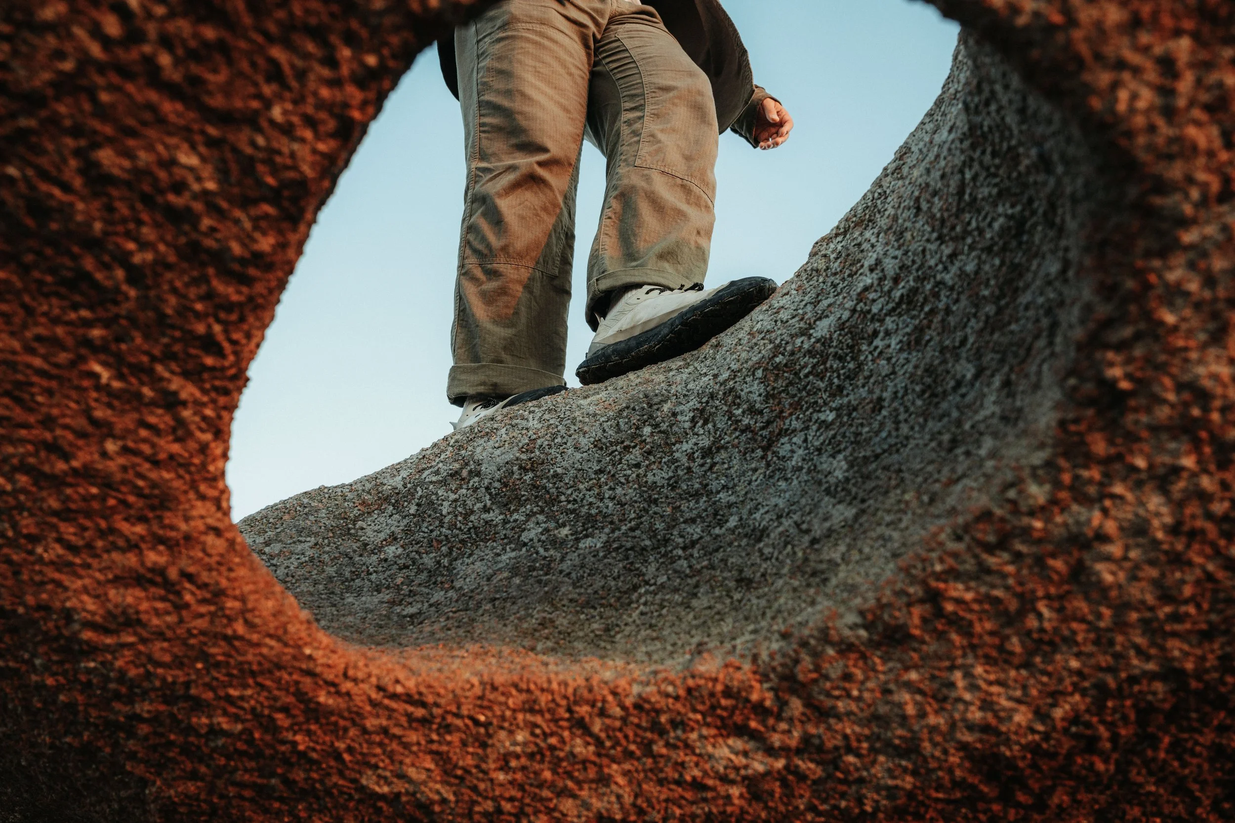 Person wearing tan pants and white shoes standing on a large rock, photographed from below through a circular opening in the rock.
