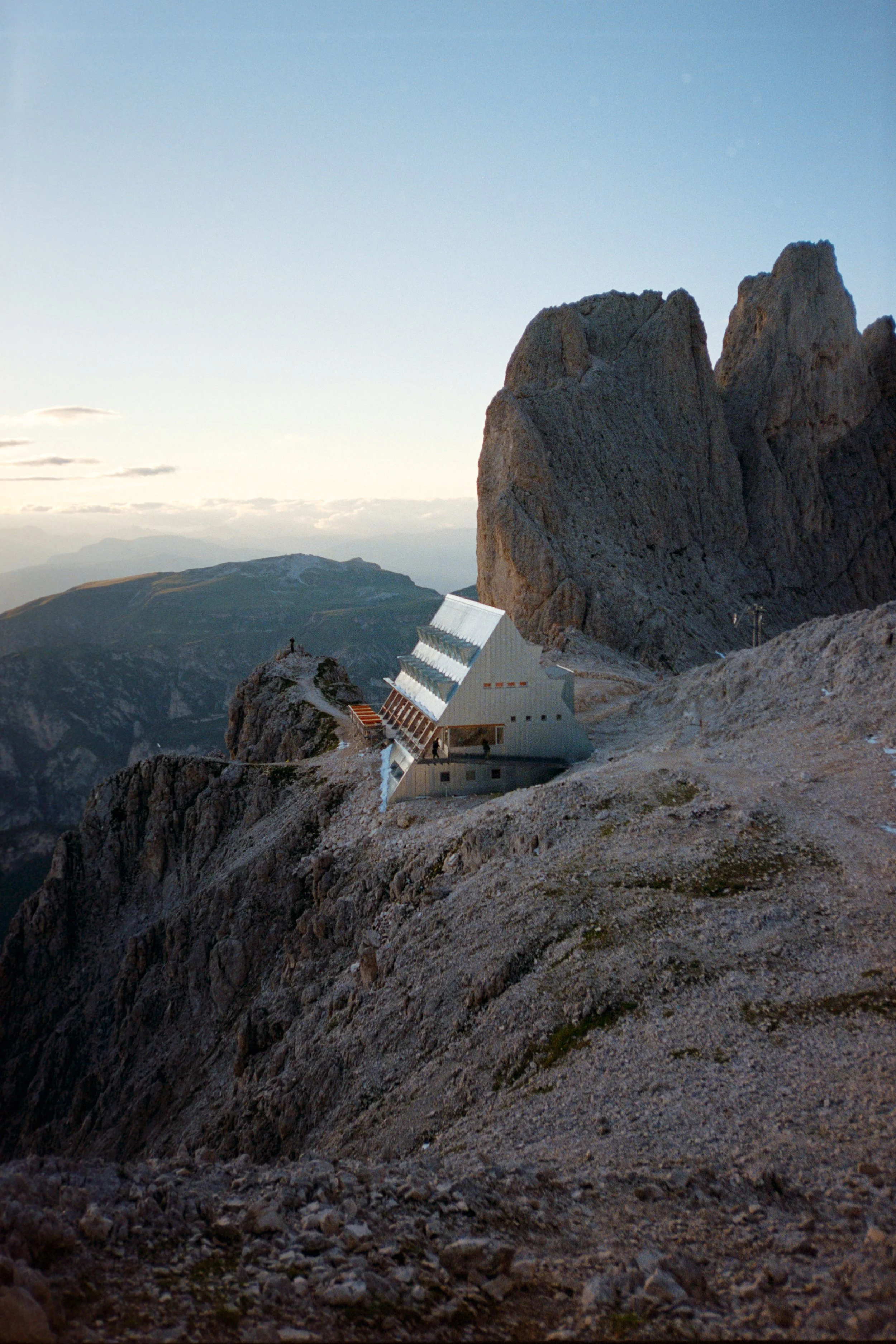 A mountain landscape with rocky terrain and a building built into the slope, surrounded by large rock formations against a faintly cloudy sky.