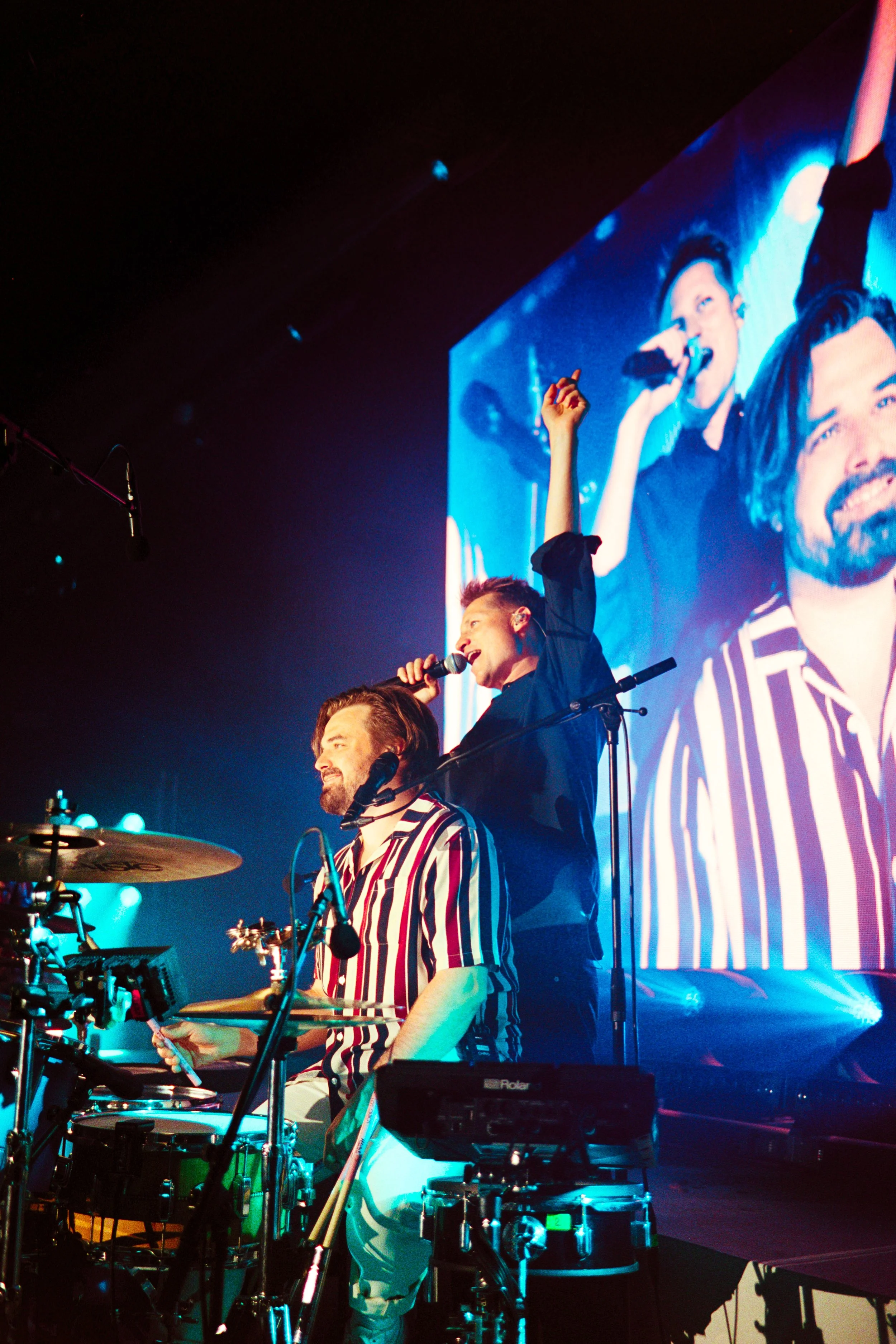 Two musicians performing on stage with one singing into a microphone and the other playing drums, with a large screen behind displaying their images.