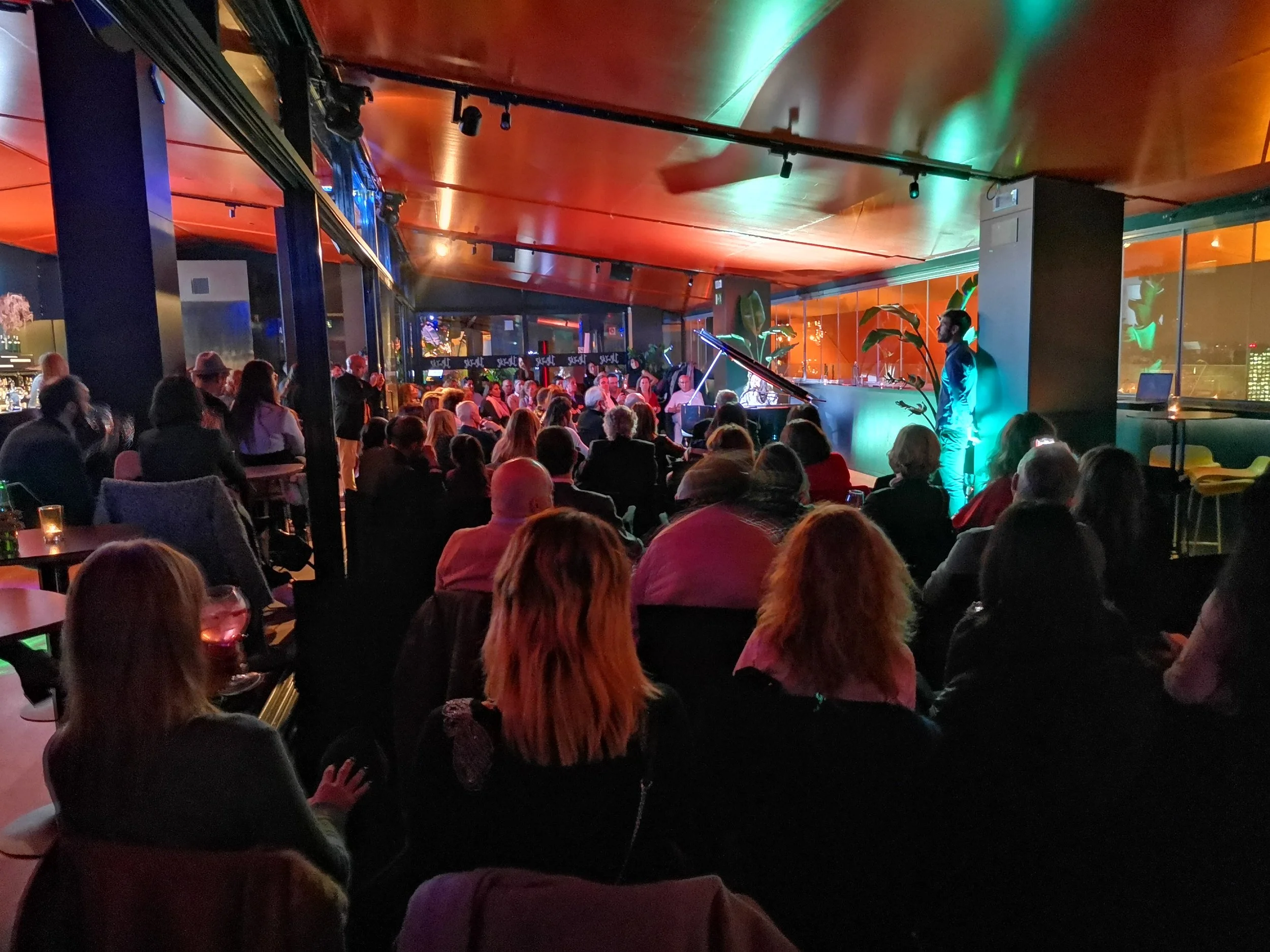 Una multitud de personas en un espacio interior, viendo una presentación en vivo de música de piano, con iluminación de colores y decoraciones modernas.