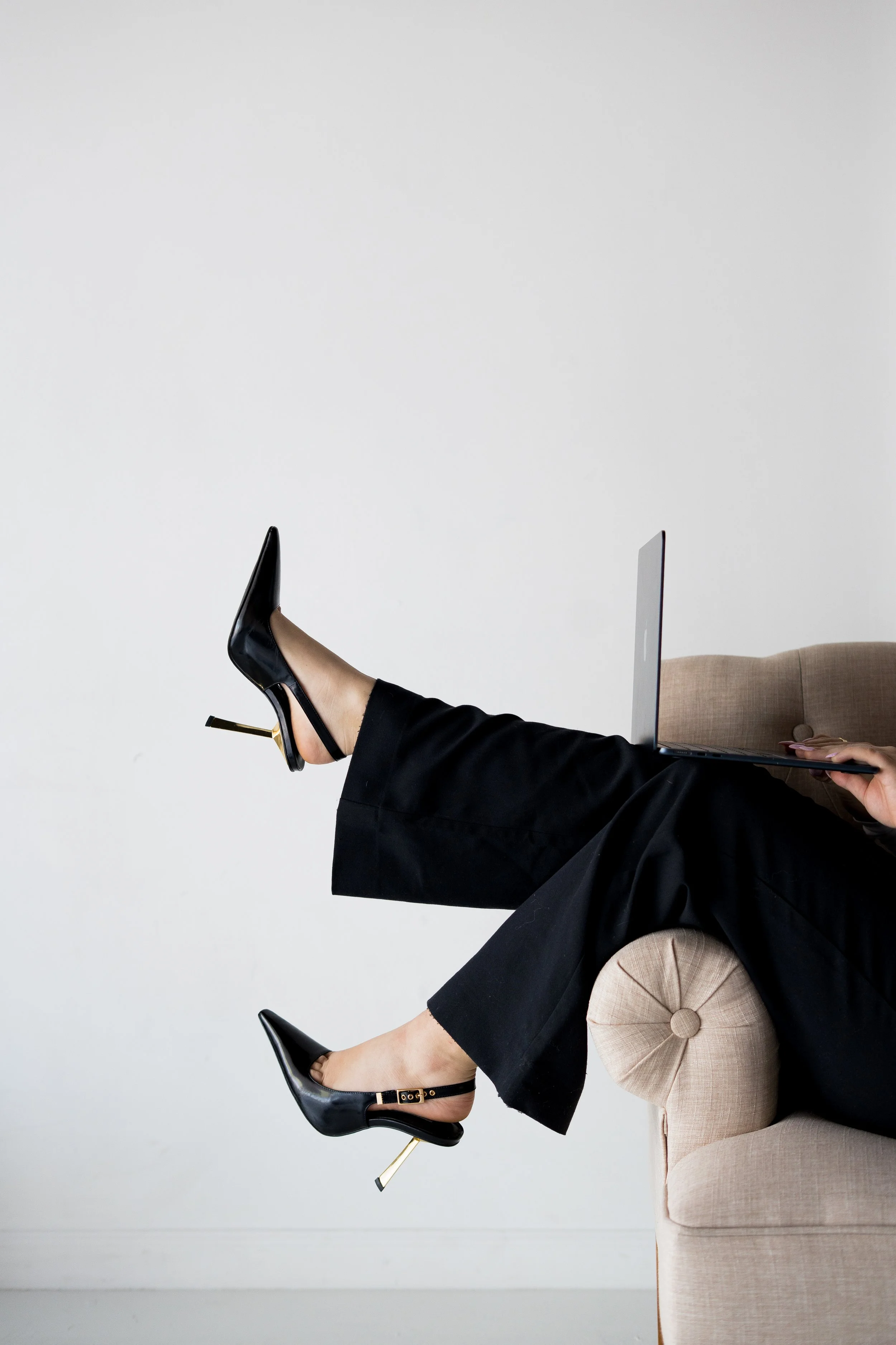 Person sitting on a beige sofa with legs extended, wearing black high-heeled shoes, holding a laptop on their lap, against a white wall background.
