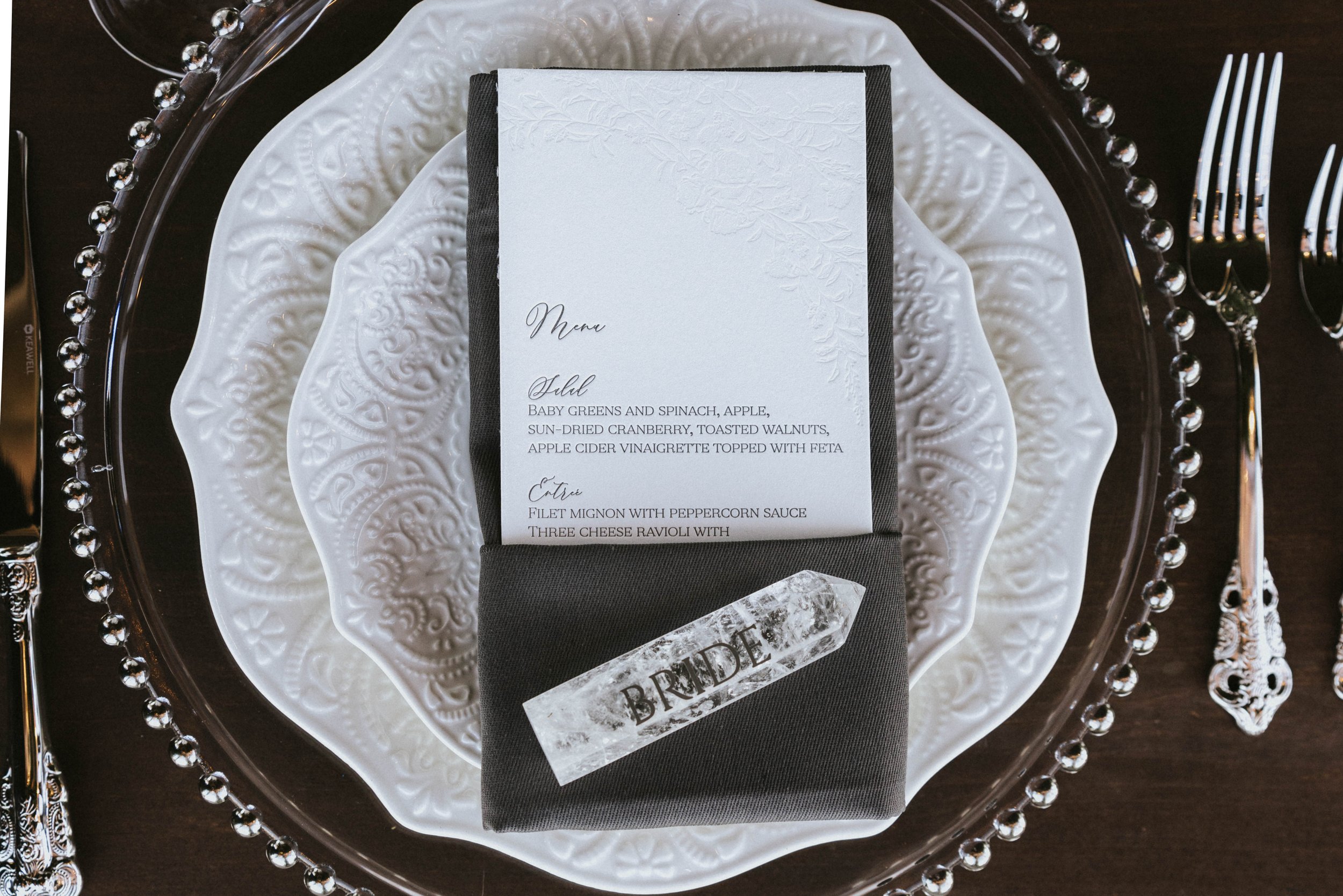 Wedding table setting with a white decorative plate, a black napkin with a translucent 'BRIDE' banner, and a menu on top, with silverware on the side.