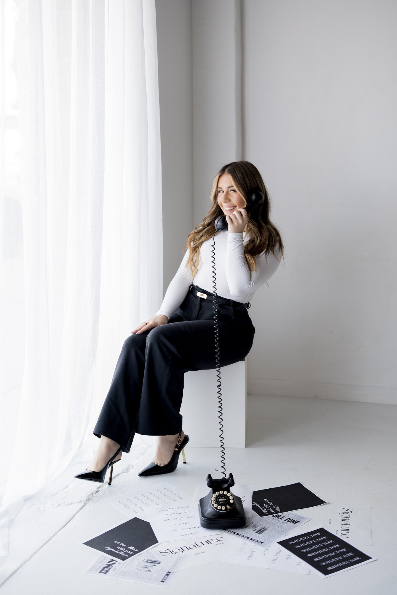 A woman sitting on a white cube, talking on a black rotary phone, with papers scattered on the floor around her, near a window with sheer white curtains.