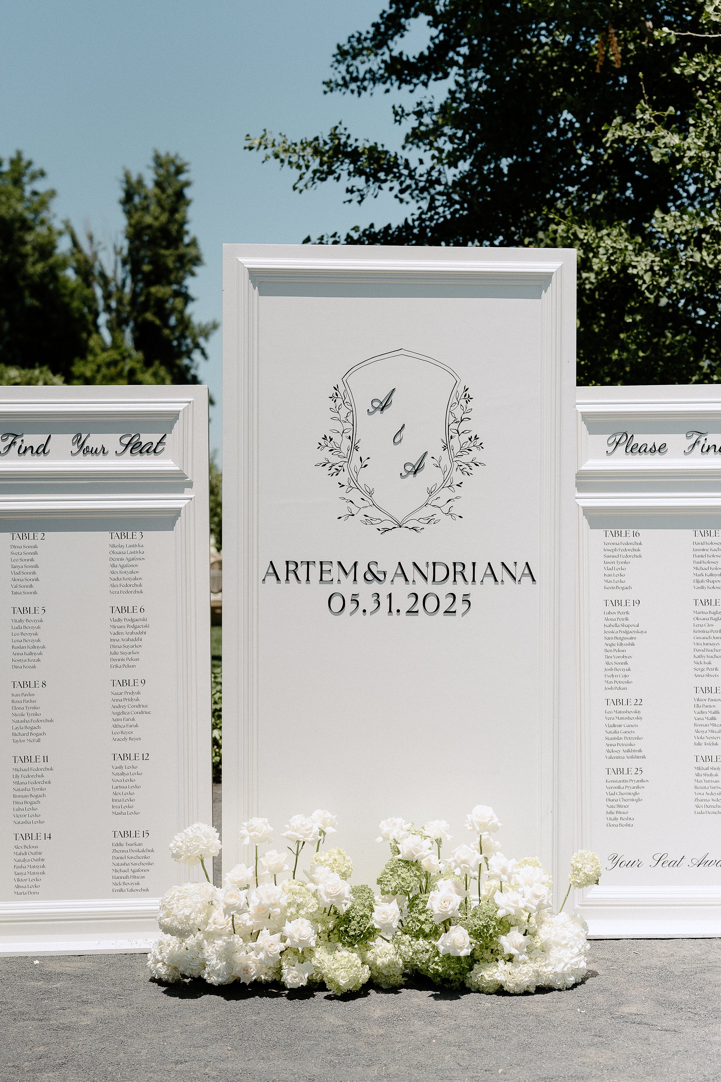 Wedding seating chart with white floral arrangements at the base, displaying the names Artem and Andriana and the date 05.31.2025, with a background of trees and a blue sky.