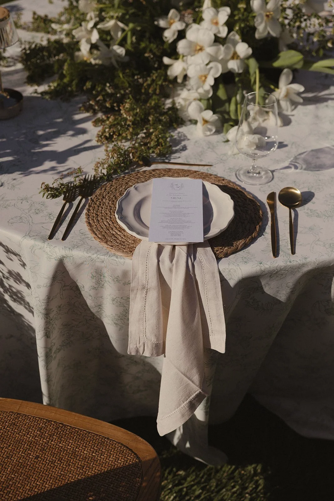An elegantly set outdoor dining table with white and gold utensils, a white plate with a menu on top, a clear water glass, and a floral centerpiece with white flowers and greenery. Elegant outdoor dining table reflecting signature tone styling, compa