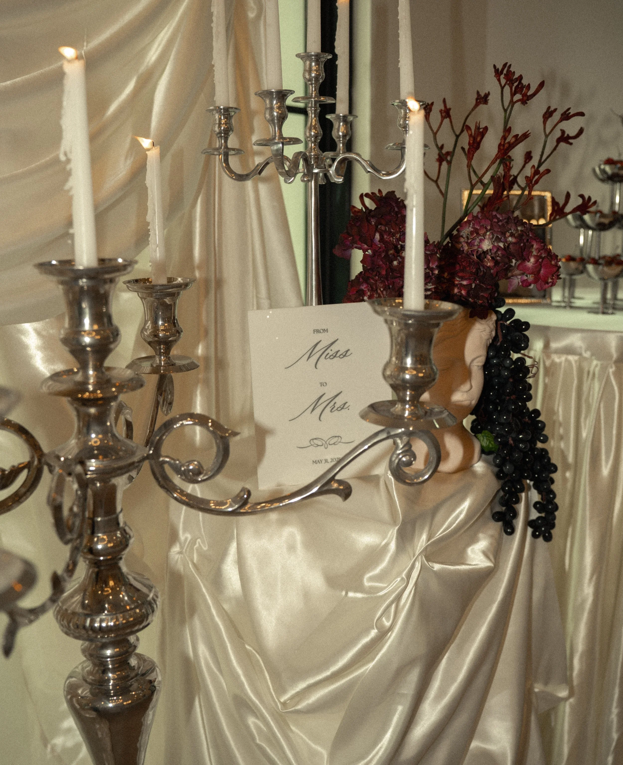 Wedding decor with silver candelabras, a card reading 'From Miss to Mrs.', a floral arrangement of red and purple flowers, and draped cream-colored satin fabric.