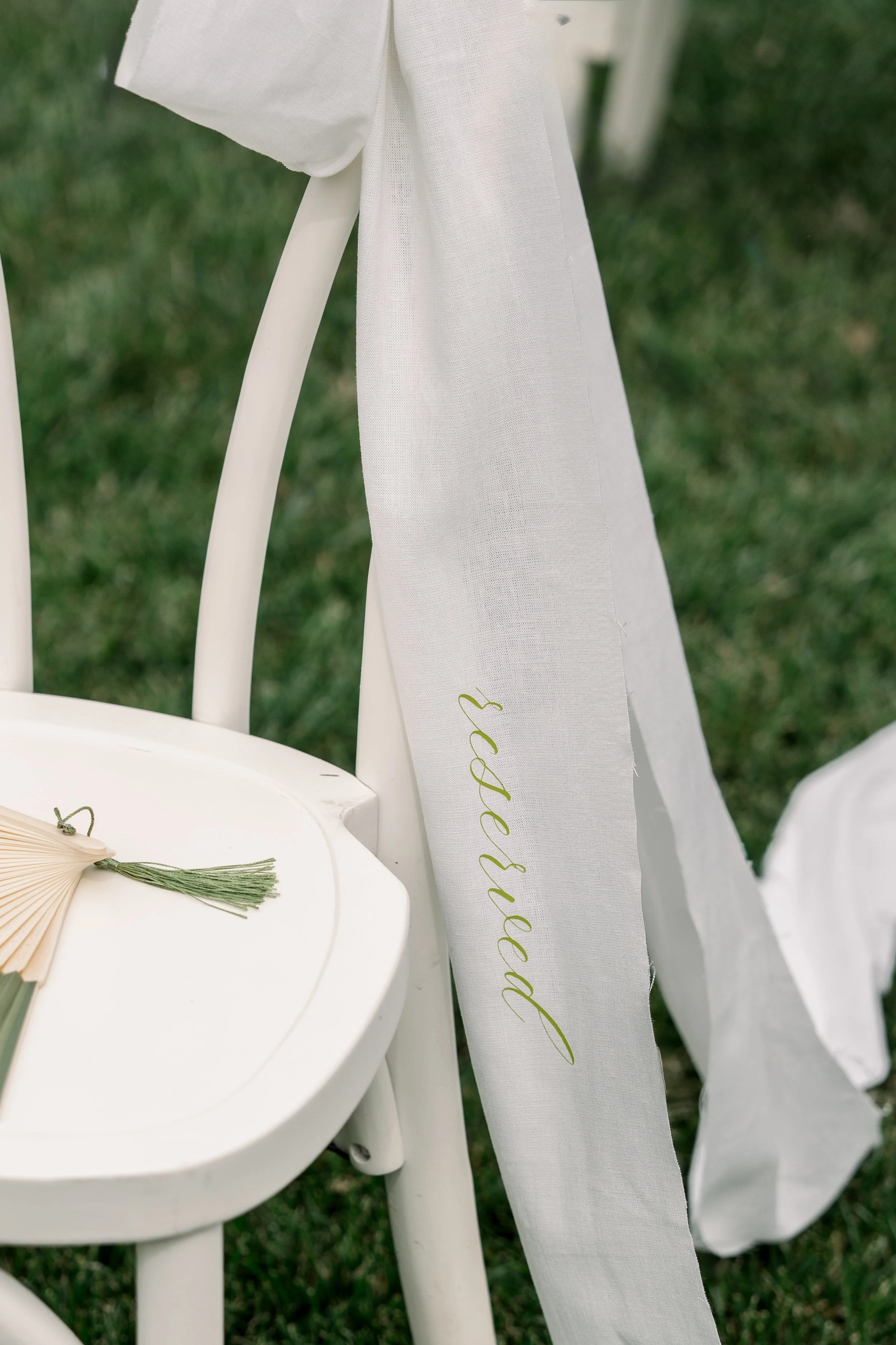 A white chair decorated with a white ribbon that has the word 'reserved' written in light green cursive, set outdoors on a grassy area.
