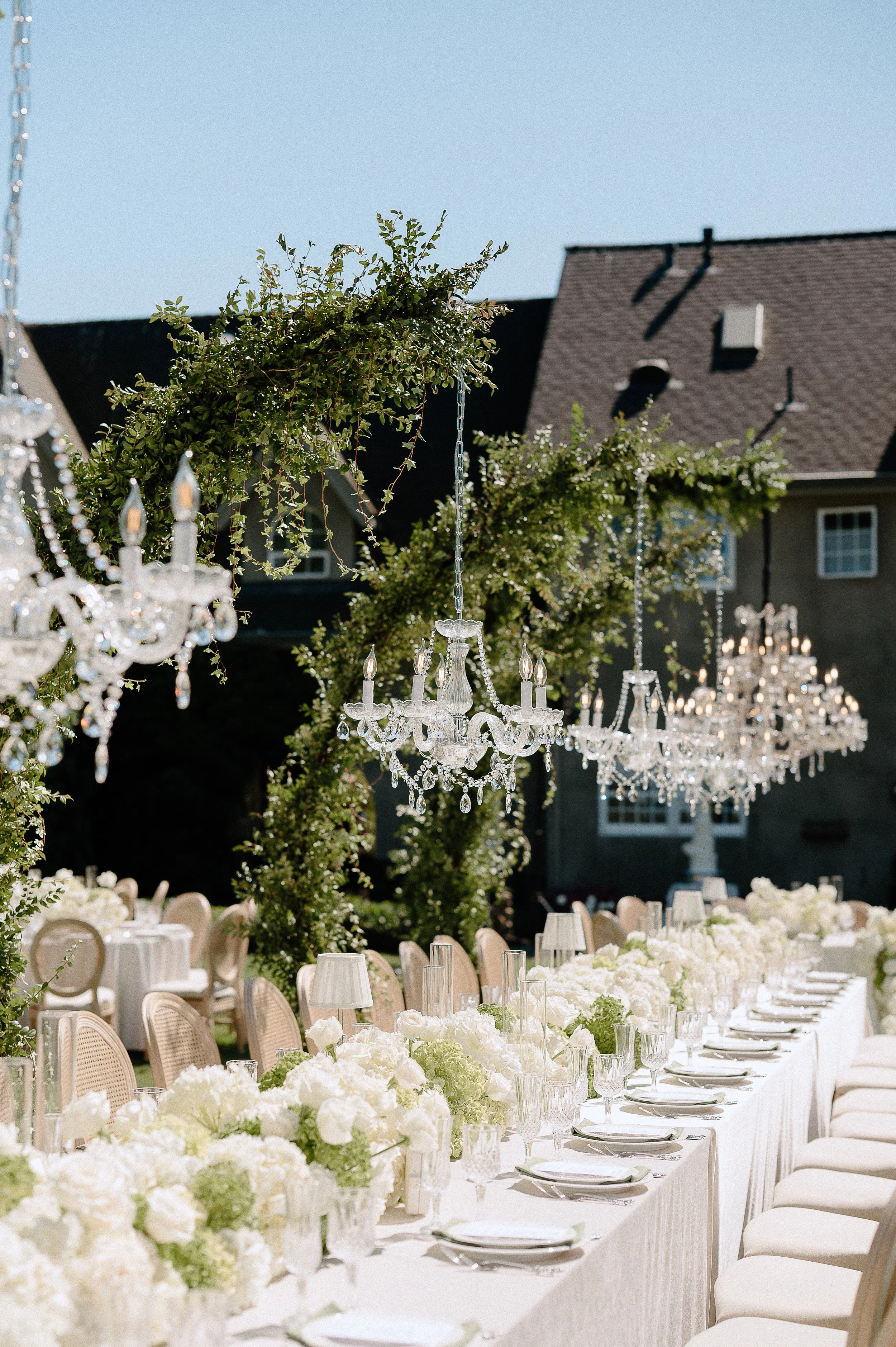 Elegant outdoor wedding reception with a long table decorated with white flowers, crystal chandeliers hanging from green archways, and a historic building in the background.