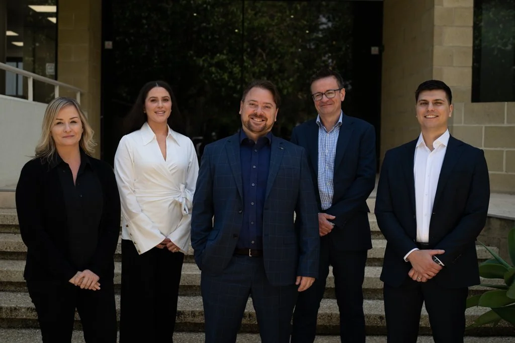 Five professionally dressed people standing on steps outside a building, smiling at the camera.