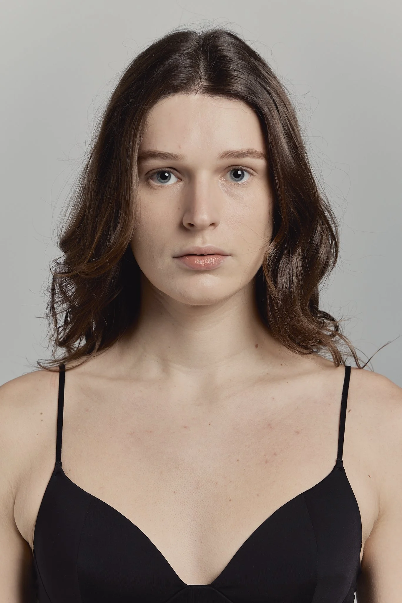 Retrato de una mujer joven con cabello castaño y ojos claros, usando un vestido negro con tirantes delgados, frente a fondo gris claro.