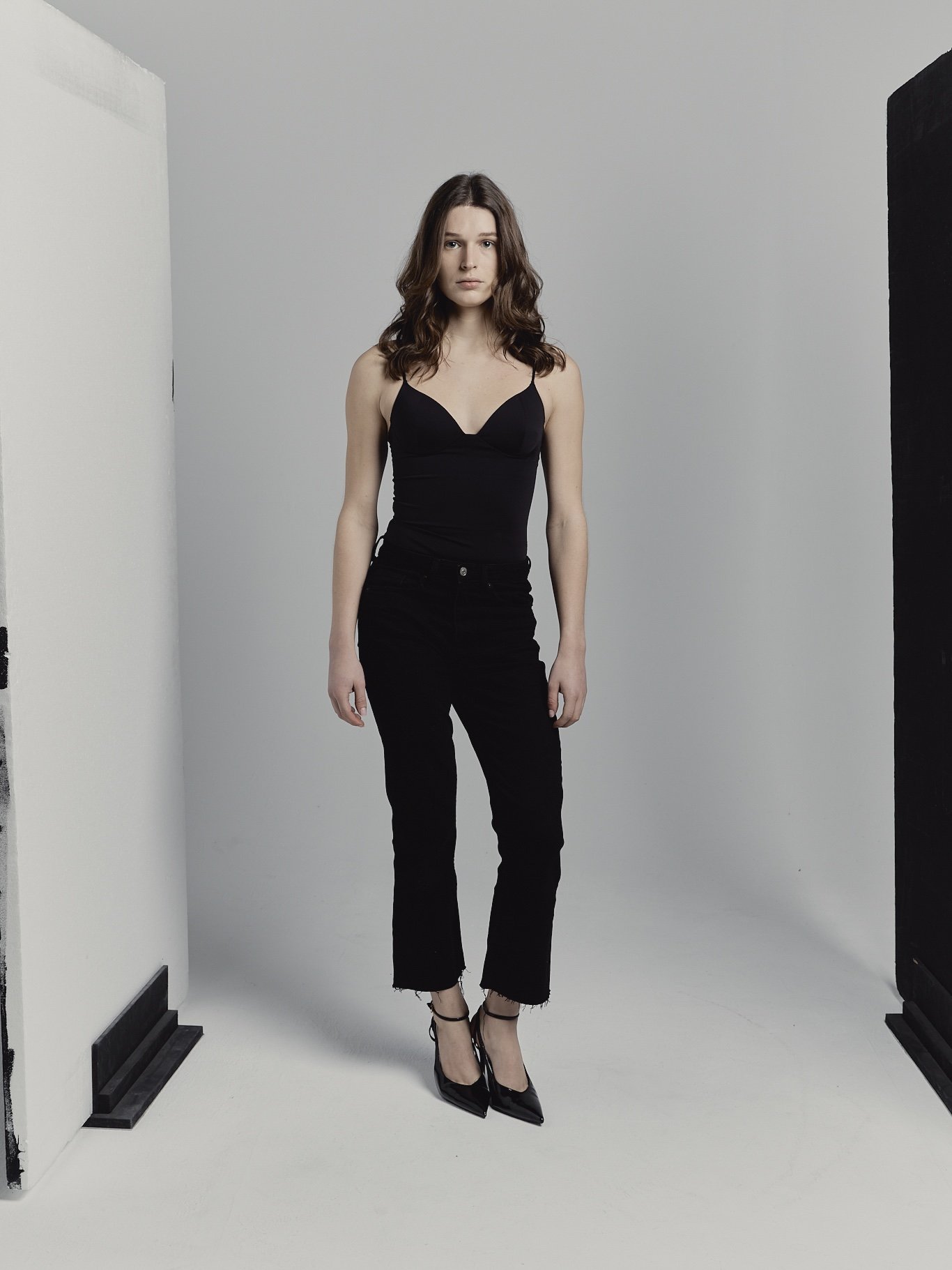 Mujer con vestido negro y tacones altos en un fondo de estudio gris con paredes blancas y negras.