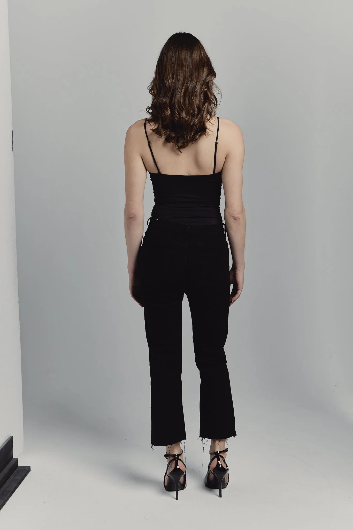 Mujer con cabello castaño oscuro y rizado, vista de espaldas, usando blusa negra ajustada y pantalones negros de tiro alto, con tacones negros de tiras.