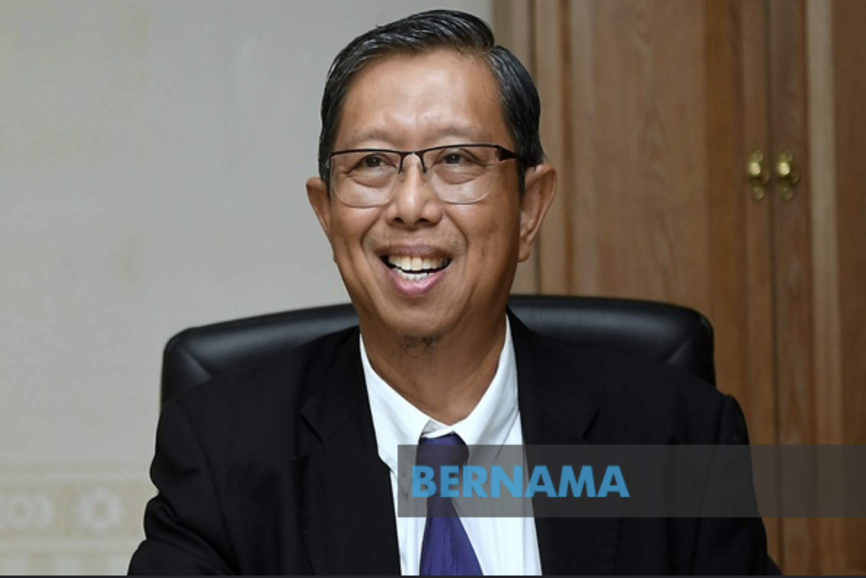 Smiling man with glasses wearing a suit and tie, sitting at a desk in an office with wooden cabinets in the background, with text 'BERNAMA' overlaid at the bottom.