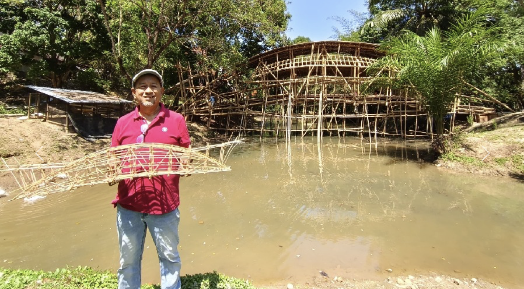 A man standing by a pond holding a bamboo model of a boat. Behind him is a bamboo structure resembling a building under construction, with trees and lush greenery around.