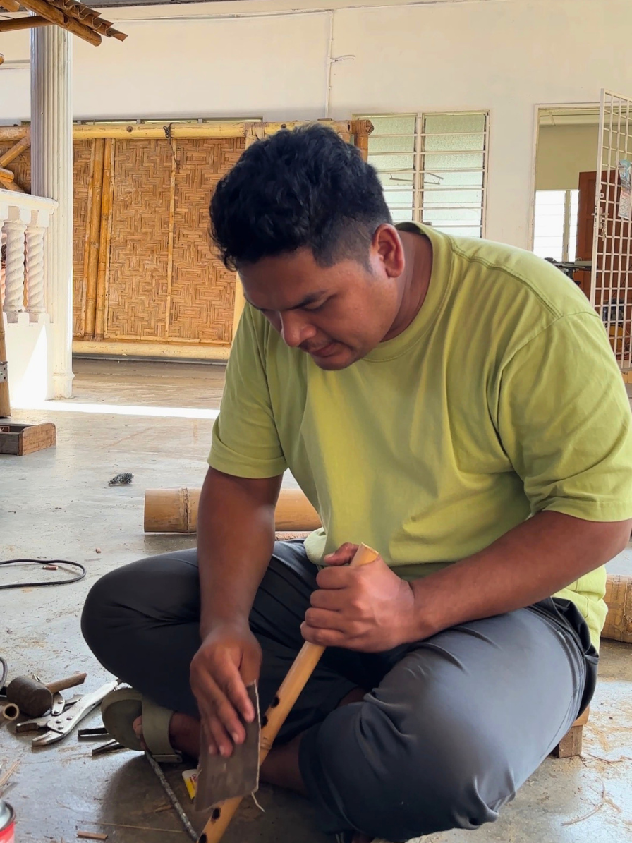 A man sitting on the floor working with a small axe on a piece of wood, surrounded by tools in a workshop or craft space.