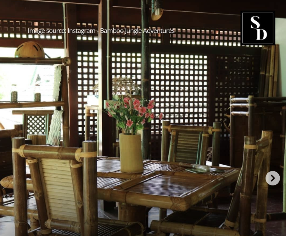 Inside of a bamboo-themed restaurant with a round bamboo table, matching bamboo chairs, and a yellow vase with pink flowers on the table. Wooden lattice walls and shelves are visible in the background.