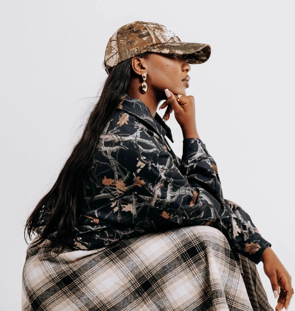 A woman wearing a camouflage cap, earrings, a camouflage jacket, and plaid pants, sitting in a thoughtful pose against a plain background.