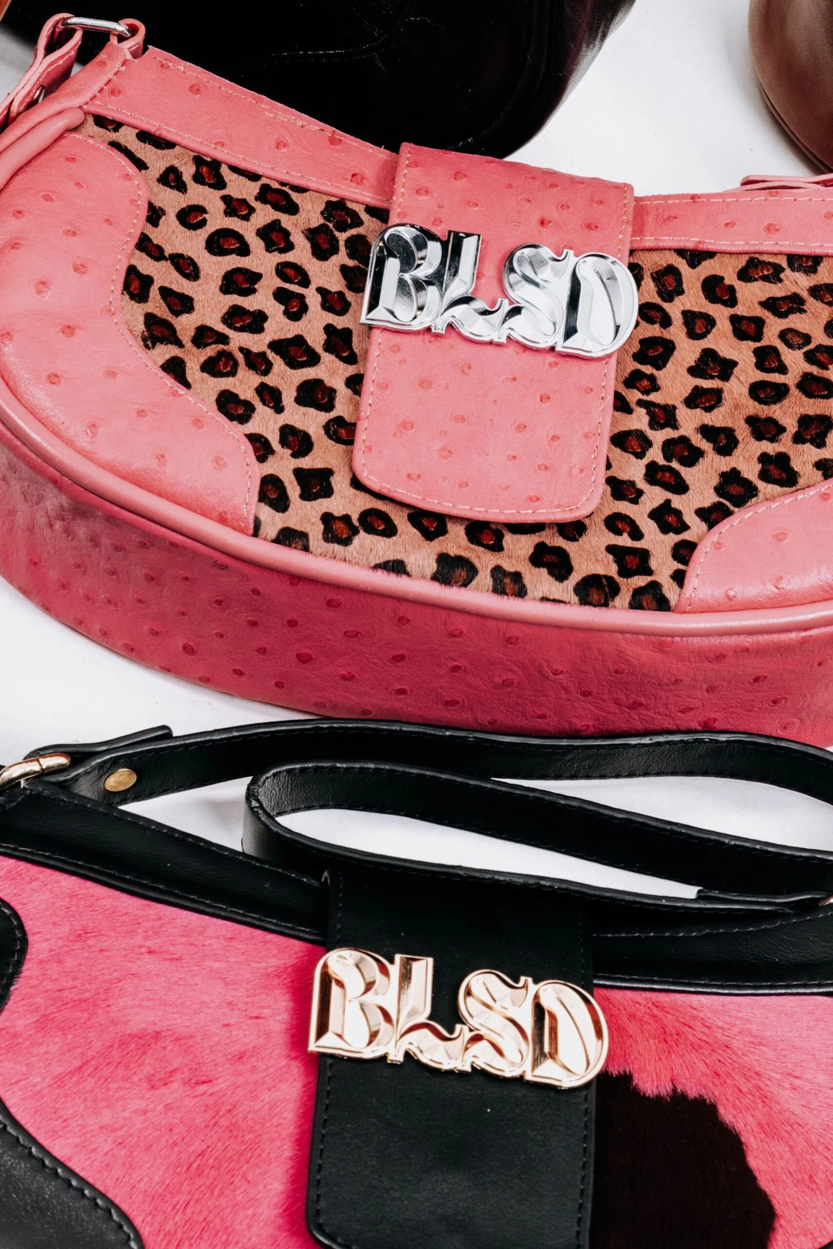 Close-up of two designer bags with bold letter hardware, one pink with a leopard print pattern and a silver 'BLSD' logo, and one pink and black with a similar 'BLSD' logo.