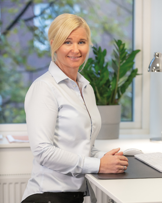 En kvinna med blont hår och vit skjorta står vid ett skrivbord i ett kontor, bakom henne finns ett fönster med utsikt över träd och en stor grönt växt.